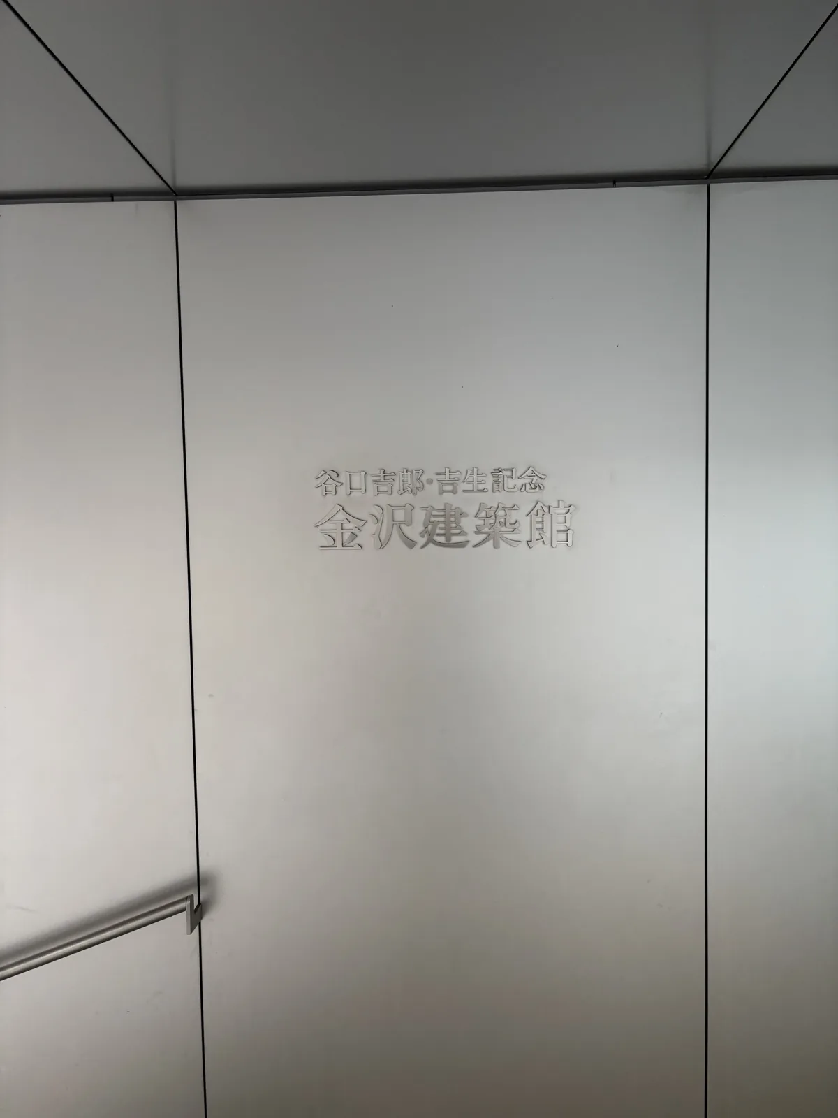 館名牆，淺灰色的金屬面板以細縫分割，中央以銀色的字刻著館名，左下方有一段扶手的端部