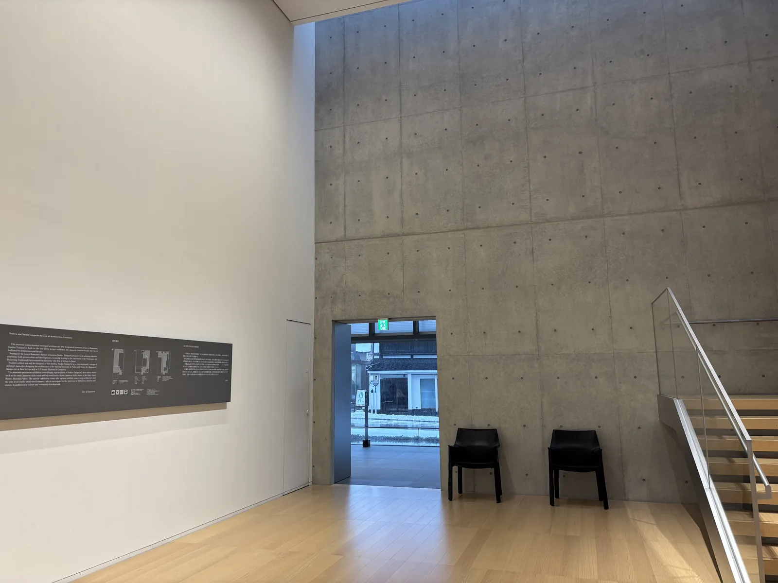 展示樓層的大廳，左側是白色的牆面與一塊深色的說明面板，右側是一整面清水混凝土牆，牆面上可見規則排列的模板孔，牆的上方有一道細長的天窗光，混凝土牆下方開著一個出入口，門外可見街屋，出入口旁放著兩張黑色的椅子，右下方是一段玻璃扶手的樓梯
