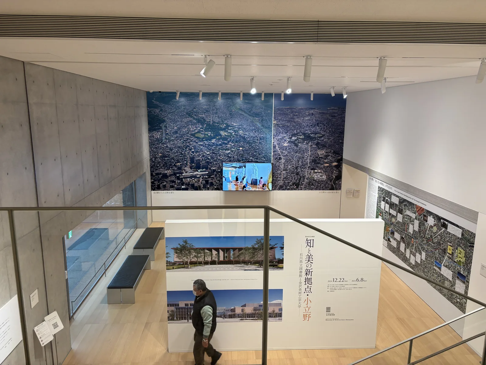 自大廳的挑空俯瞰地下企劃展示室，前景是玻璃扶手，下方是木地板的展示空間，正面的牆上是一幅巨大的空拍照片與一台螢幕，中央立著一面展覽的標題牆，左側是清水混凝土牆與一排長凳，右側牆上貼著地圖