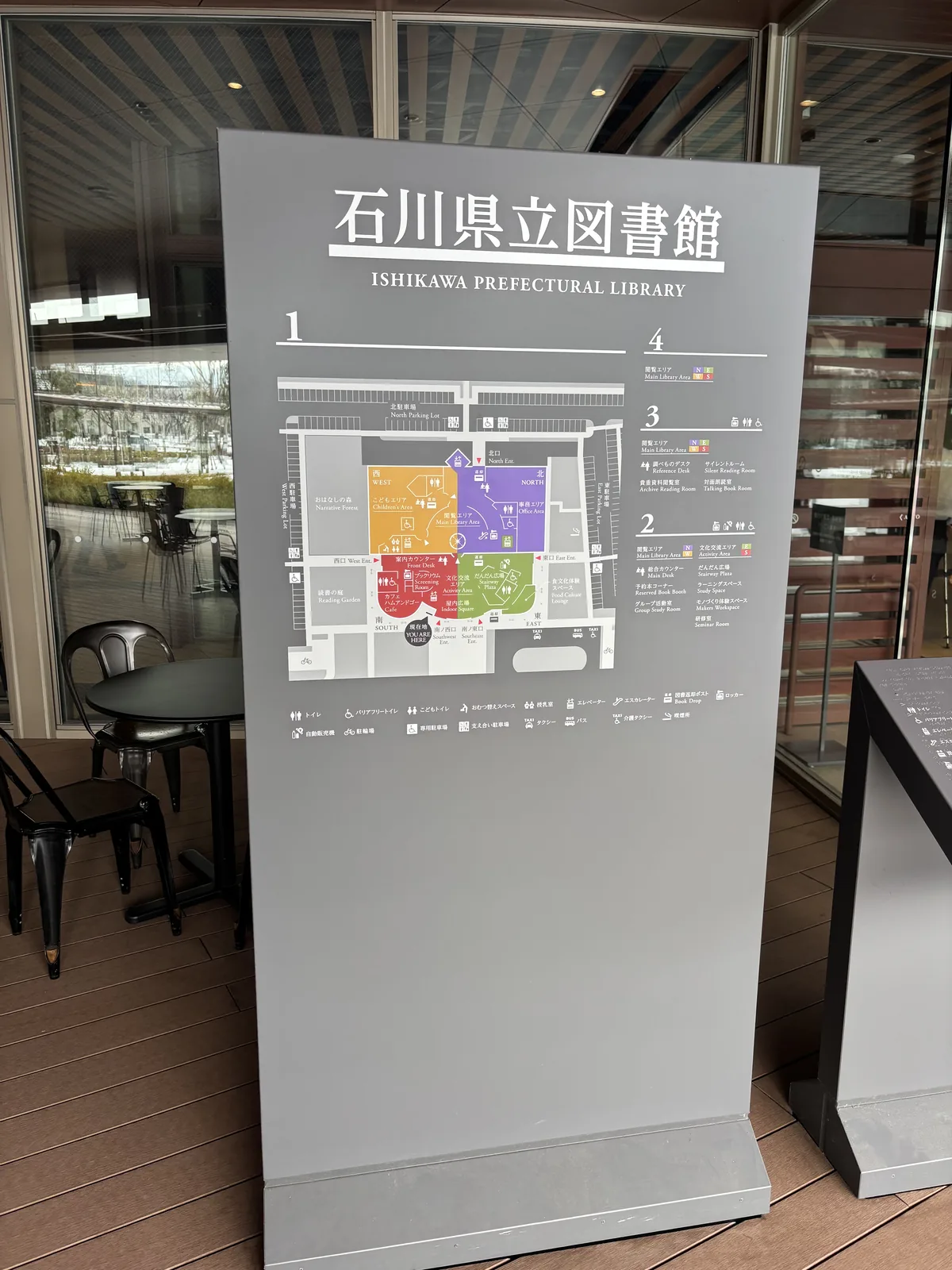 館名看板，淺灰色的立牌上方寫著館名與英文名，下方是一張以彩色區塊標示各樓層的平面圖與圖例，立牌後方是玻璃牆與屋外的桌椅