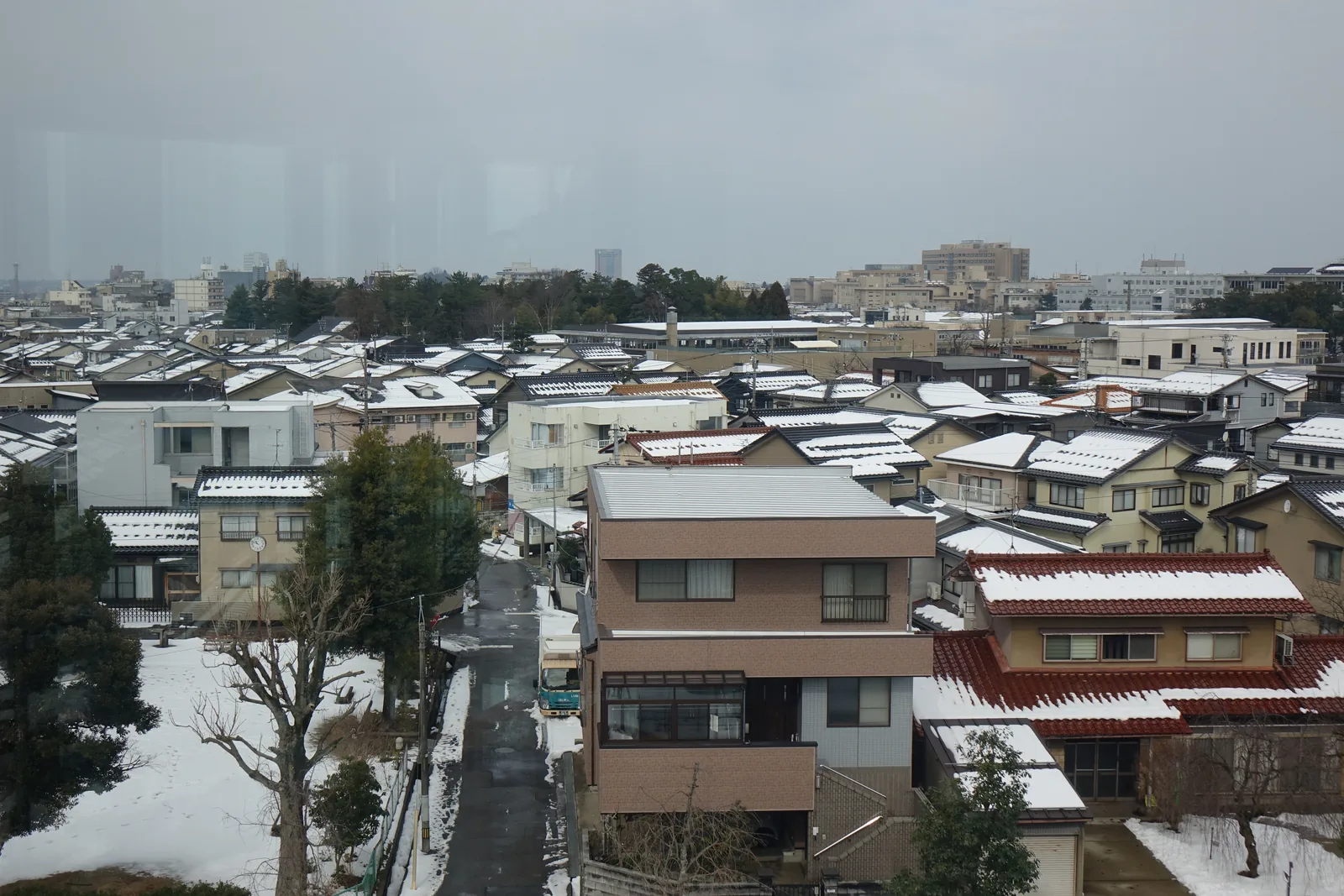 自圖書館上層的窗戶望向北側，窗外是一片覆雪的住宅街，屋頂上積著白雪，近處有幾棵樹與一棟淺色的公寓，遠處是低矮的丘陵與陰沉的天空