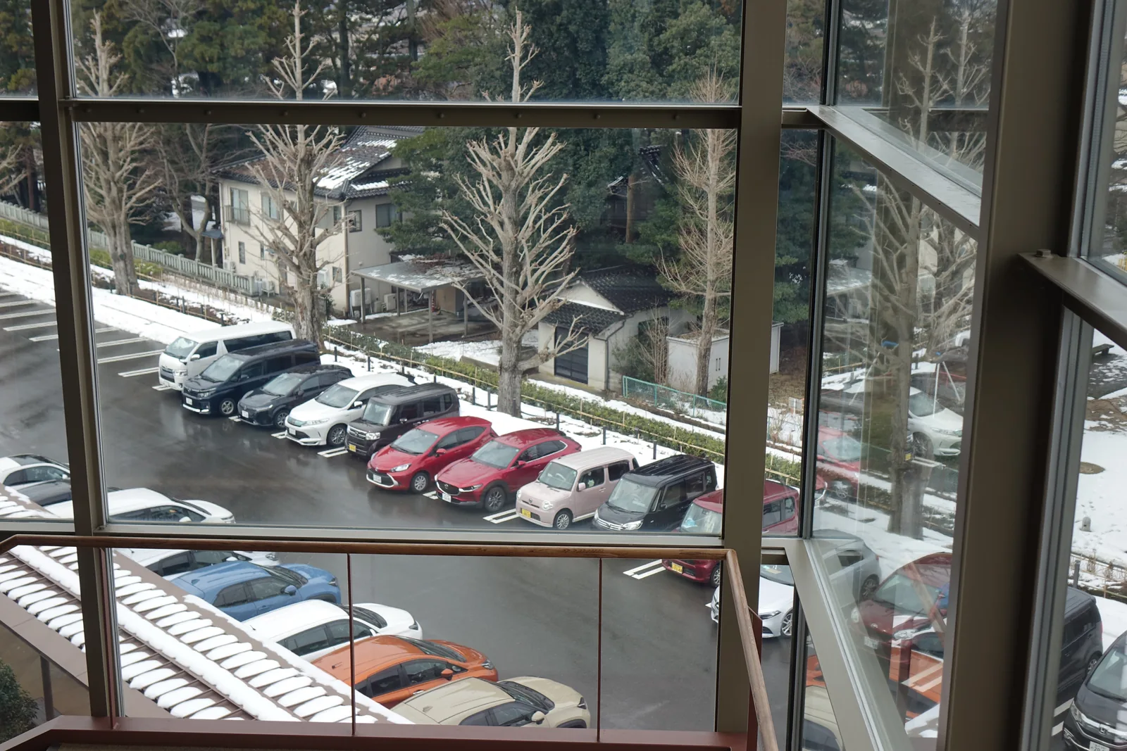 自圖書館上層的窗戶俯瞰停車場，玻璃與金屬框之外是一片停滿汽車的停車場，周圍種著一排落葉的樹木，地面殘留著積雪，前景是玻璃扶手的上緣