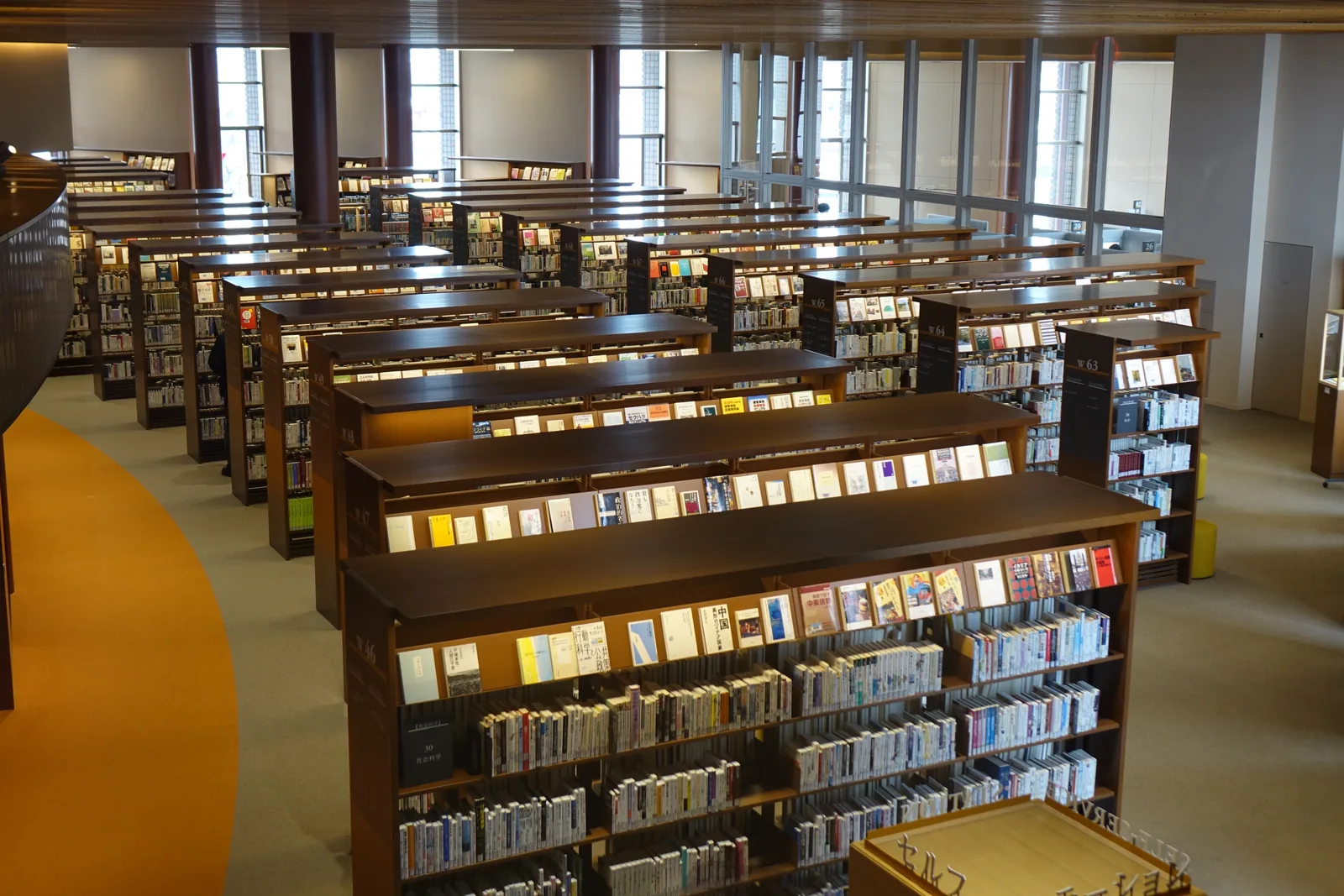 大閱覽空間外周的書架區，一排排木製書架沿著外牆整齊排列，地面一側是橙色的地毯帶，遠處是高窗與明亮的外光，上方是格柵狀的天花