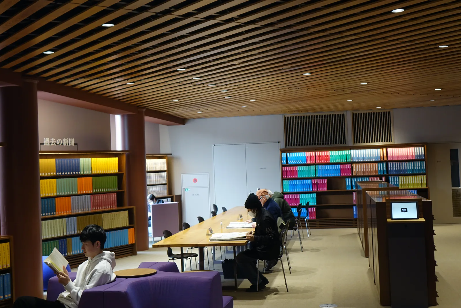 一間閱覽室，左右兩側是排滿彩色書脊的書架，中央是一張長條的閱覽桌與幾位讀者，前景是一張紫色的沙發與一位正在看書的讀者，天花是木條格柵