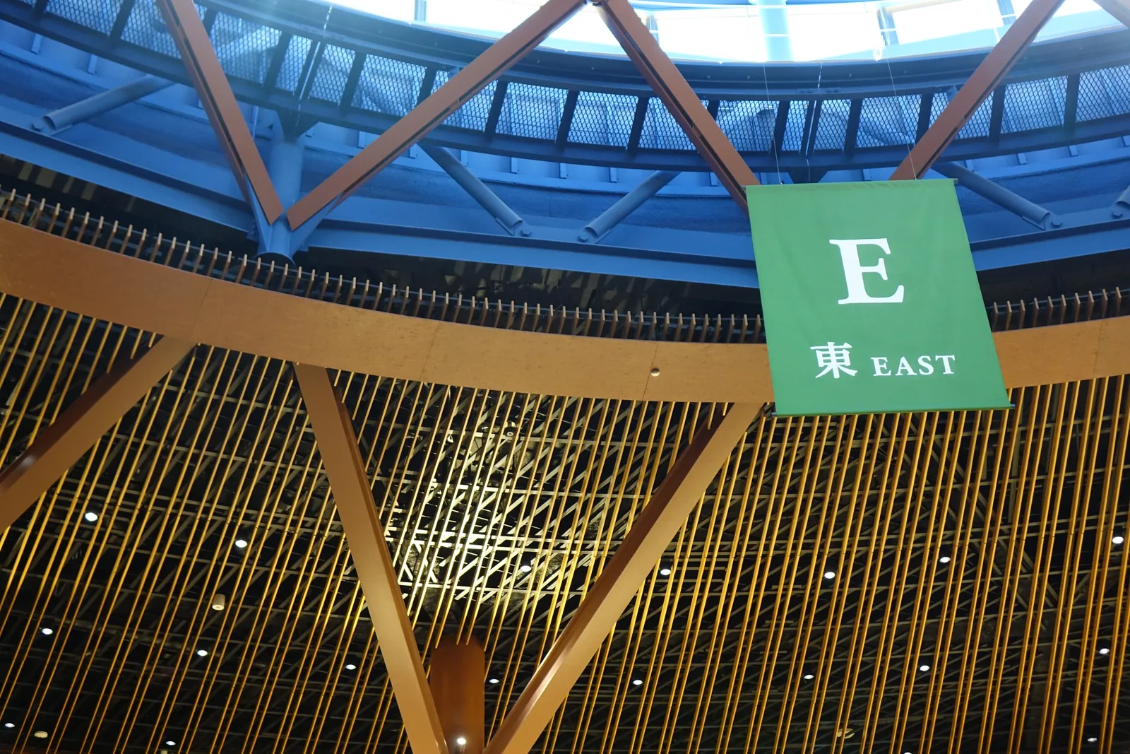四樓回廊的一角，一面綠色的旗幟寫著「E」與東，垂在紅褐色的斜向構件之前，構件之後是藍色的天花與白色的高窗，下方是層層的木條天花