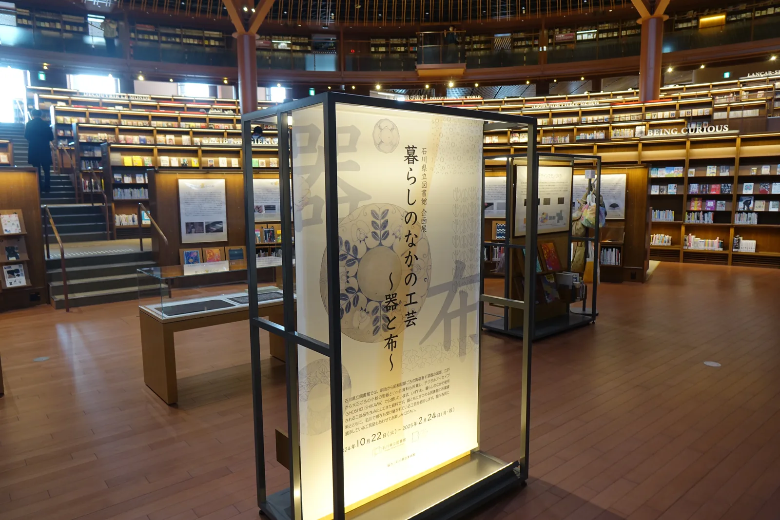 一樓的展示區，前景是一座以玻璃與金屬框構成的展示看板，看板上寫著企劃展的標題，後方是展示櫃與書架，地面是深色的木地板，遠處是通往上層的階梯