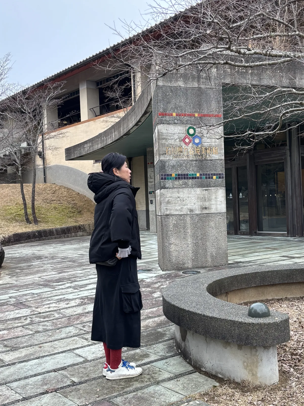 美術館入口前的廣場，一根以混凝土砌成的方柱立在畫面中央，柱上嵌著彩色的圓形標誌與館名，右側是一段弧形的混凝土矮牆，後方是建築的入口與一道弧形的混凝土牆，地面鋪著石板，一位參觀者站在柱旁
