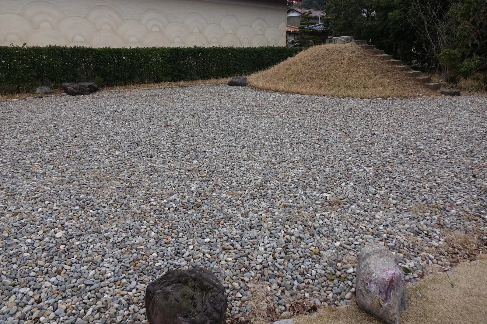 館外的一處庭園，地面鋪滿灰色的砂礫，砂礫上散置著幾塊石頭，後方是一座覆著枯草的土丘與一道修剪整齊的綠籬，土丘旁有一段石階，最遠處是一道白色的牆