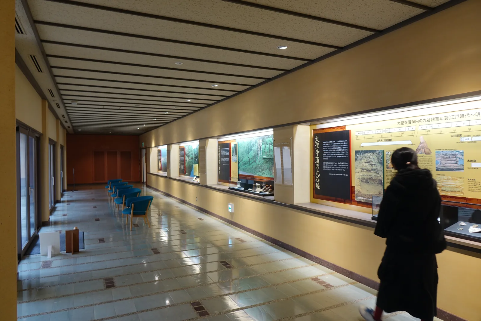 一條細長的展示廊，左側是一排落地窗與幾張藍色的椅子，右側是一整排內嵌的展示櫃，櫃內是說明板與展品，天花是淺色的平面與一列列的線條，地面是光亮的磁磚，一位參觀者站在展示櫃前