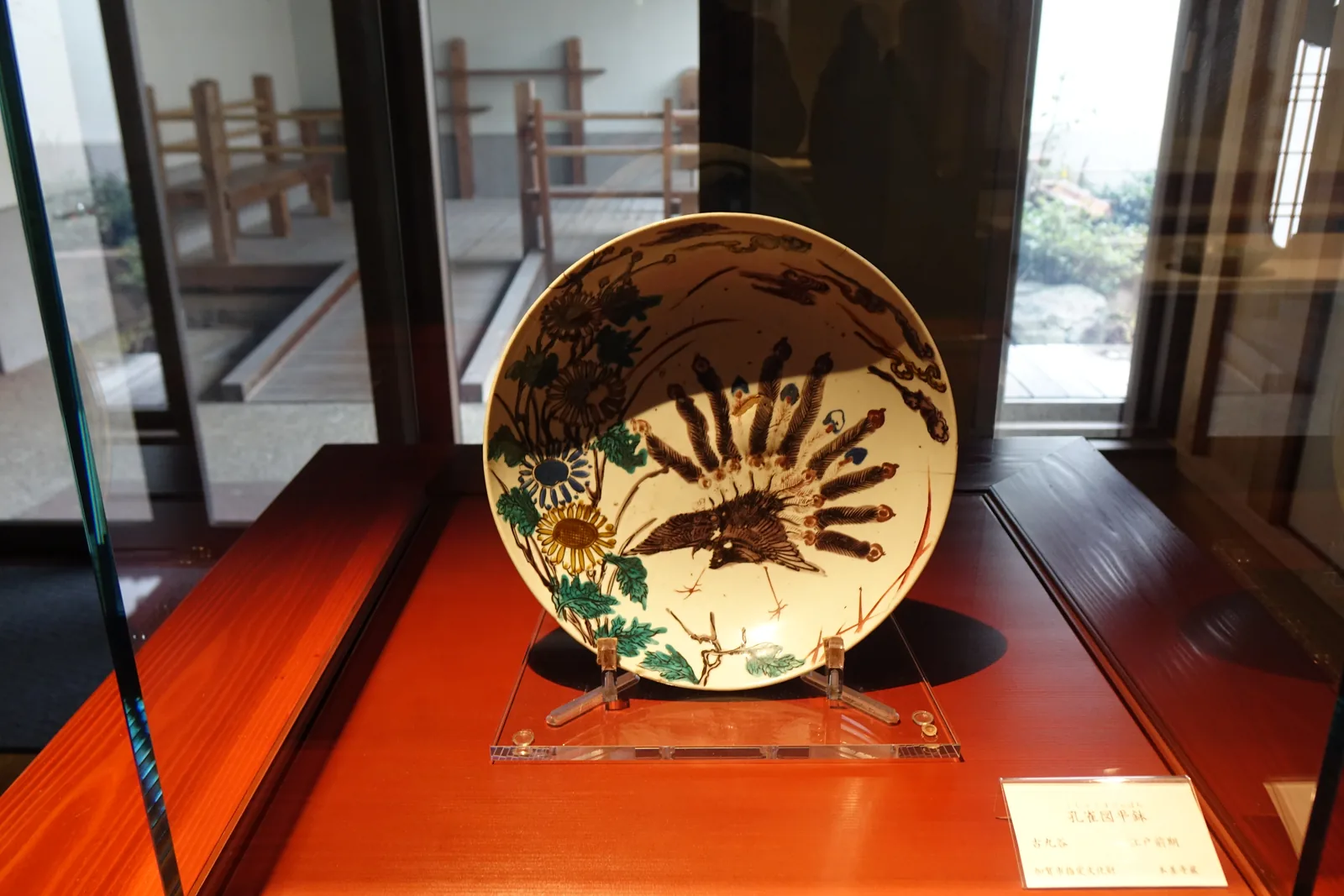 一只彩繪的大盤立在展示台上，盤面上畫著一隻黑色的鳥與綠色、黃色的花草，展示台是紅褐色的漆面，盤後方是一扇玻璃窗，窗外可見木製的柵欄與庭園