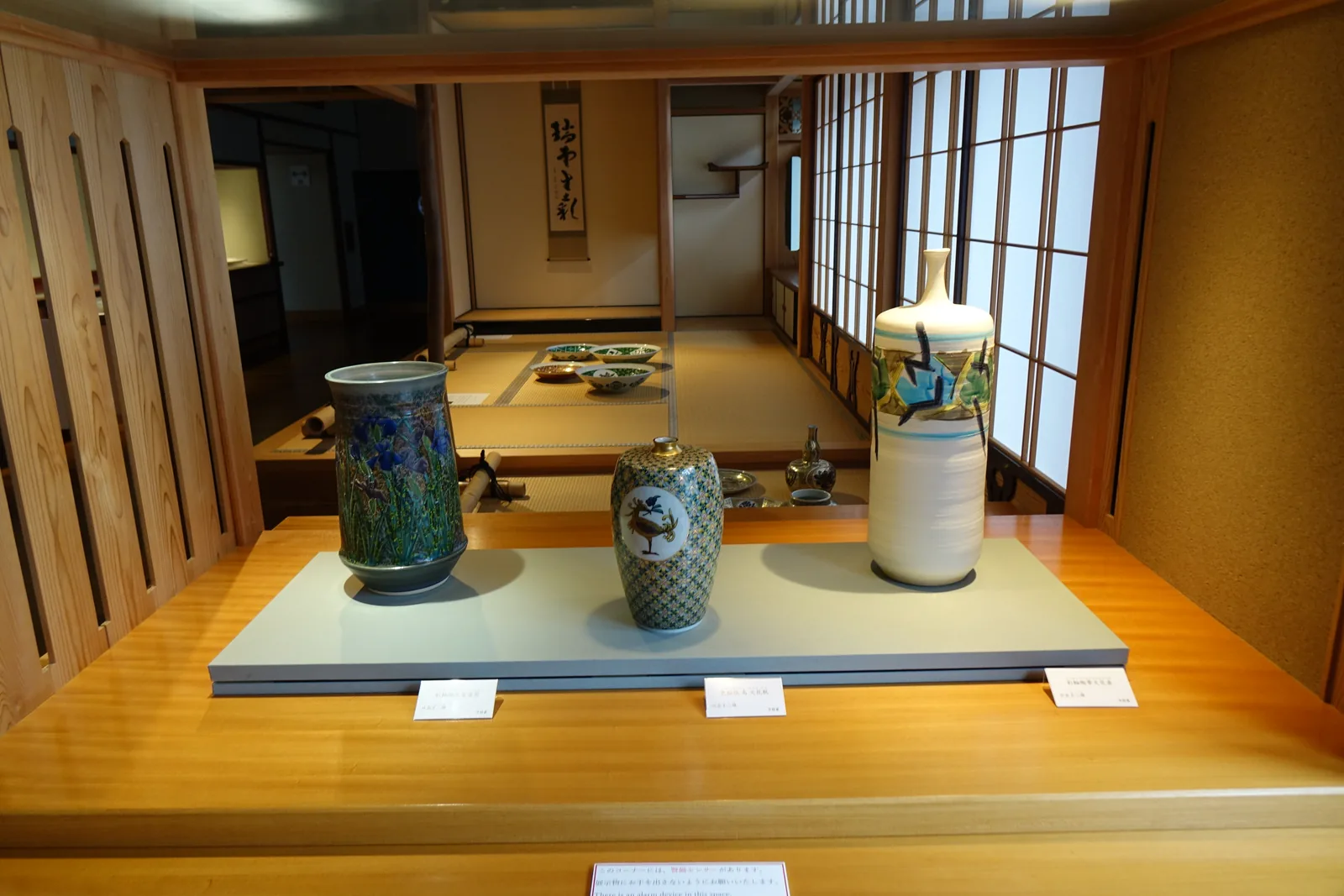 一座內嵌於木框中的展示櫃，櫃內的淺灰色展示台上排著三只壺與瓶，兩只是深綠色與藍色的彩繪，一只是白底的長頸瓶，展示櫃的後方是和室的障子與書軸，左右是木格的門扇