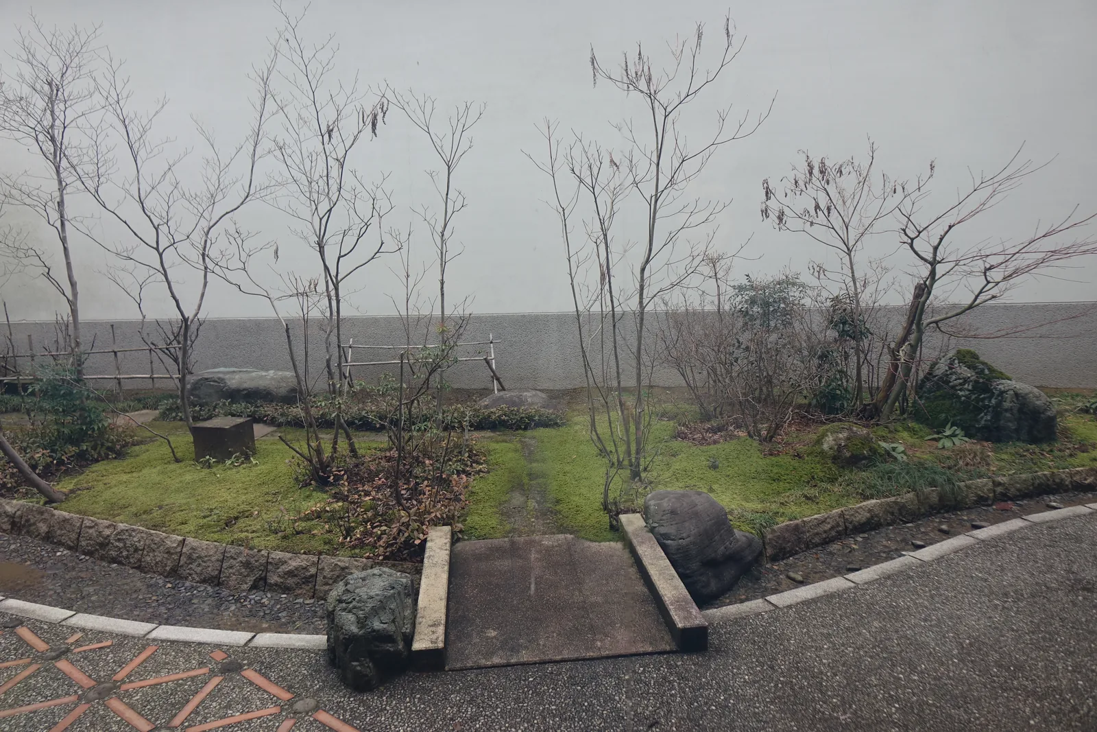 北側的庭園，以白色的牆為背景，幾棵細瘦的落葉樹立在青苔與草地上，中央是一條以兩根木樁收邊的小徑，前景是弧形的石砌邊緣與鋪著碎石的地面，地面上嵌著紅色的磚紋