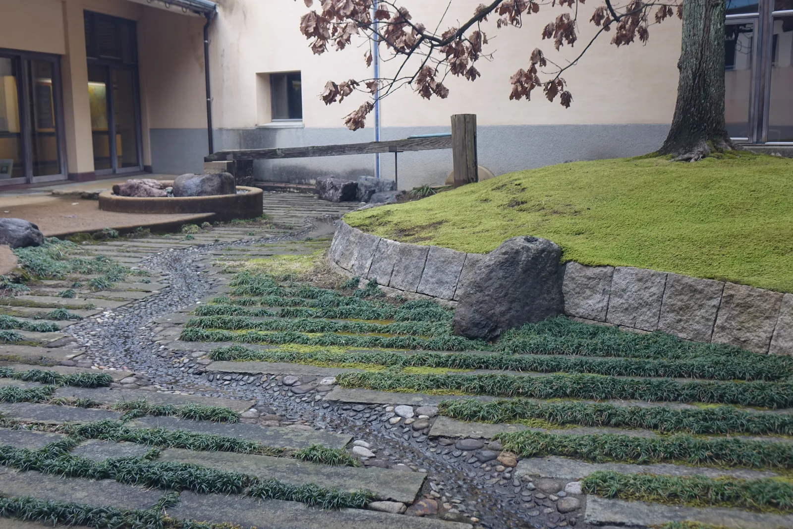 兩棟建築之間的庭園，一條以碎石鋪成的細流從右上方蜿蜒而下，細流兩側是一階一階的草帶，右側是一座以石塊砌邊的青苔丘與一塊大石，後方是建築的灰泥外牆與窗，左上方是一棵枯葉的樹