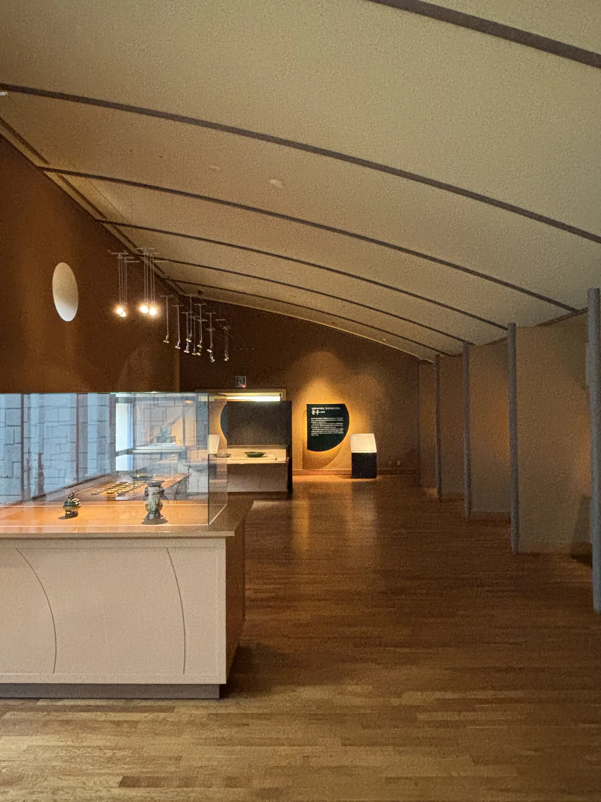 一處展示空間，天花是一道緩和的弧形曲面，左側是一座白色的展示台與玻璃展示櫃，正面的牆上有一個圓形的燈具與一幅照亮的說明板，右側是一列淺色的牆面，地面是木地板