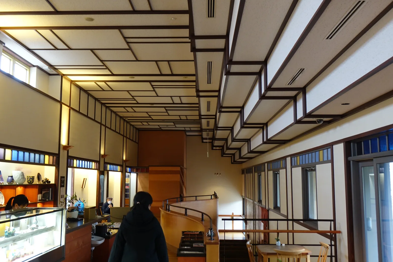二樓長廊的另一端，左側是陳列陶器的玻璃櫃與層架，右側是一道弧形的木製扶手與通往一樓的樓梯，天花是格狀的木線條，遠處是高窗，一位參觀者走在長廊中