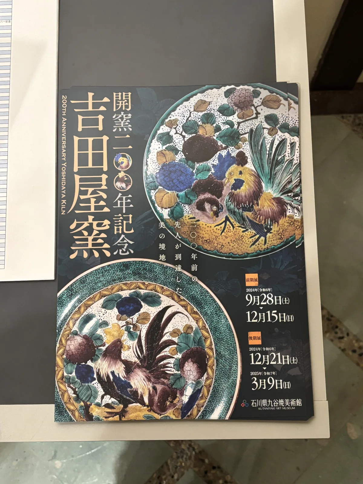 吉田屋窯特別展的海報，深色的底上印著兩只彩繪的大盤，盤面上畫著公雞、牡丹與花草，海報上方以白字寫著展覽名稱，右側是會期，海報掛在一面木製的牆上