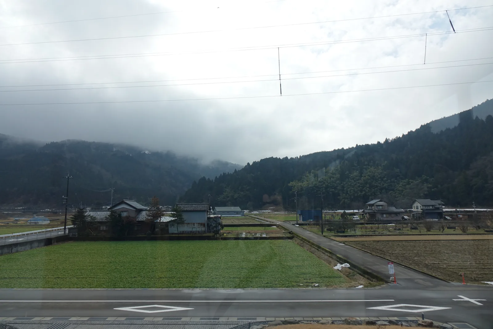 自二樓的玻璃向外望，近處是草地與道路，中景是幾棟民家與一片綠色的田，遠處的山腰以上被低垂的雲遮住
