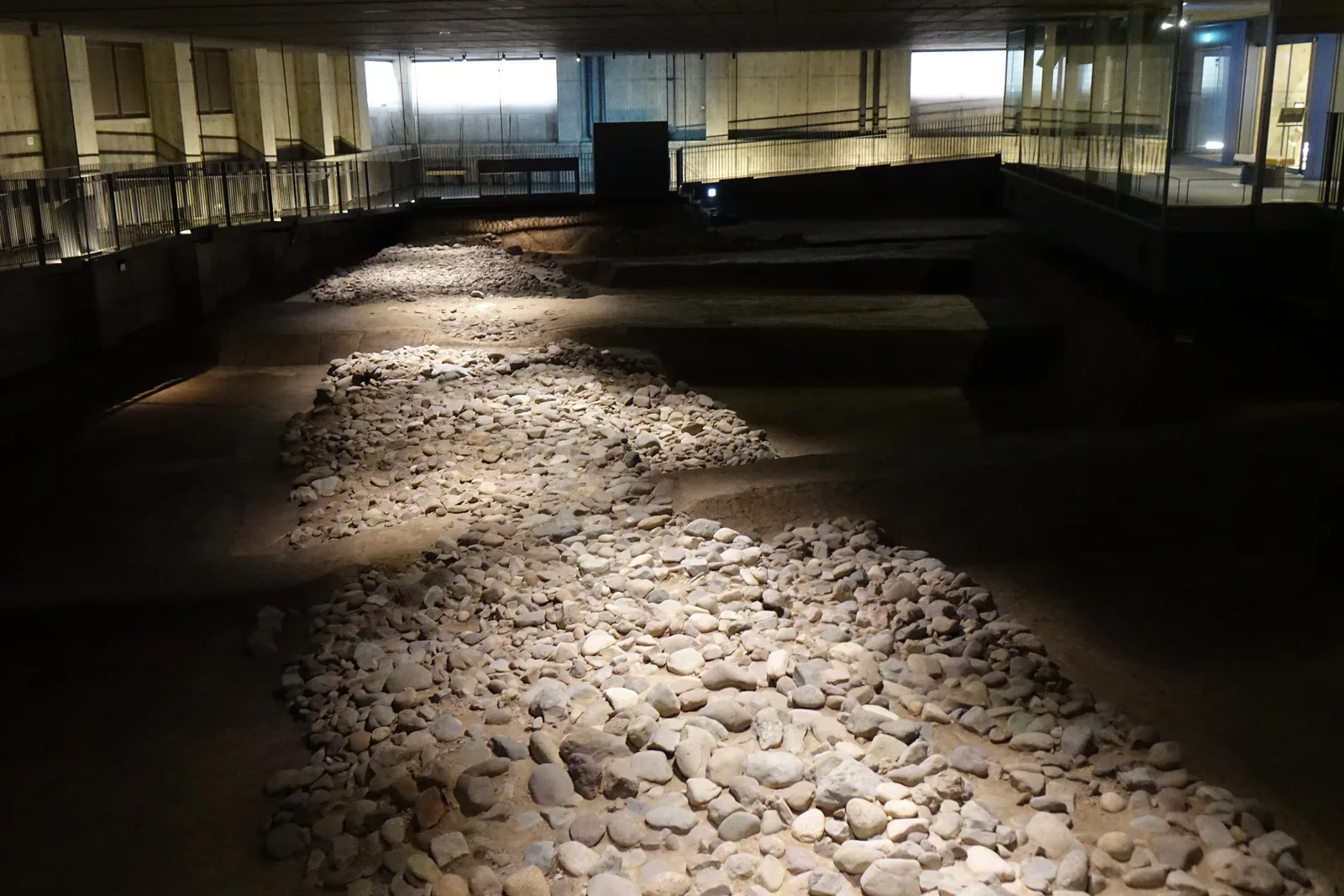 遺構展示室的內部，中央是一片以河原石鋪成的石敷，石敷在燈光下浮現，四周的地面沉在暗處，遠處是一圈設有欄杆的迴廊與幾扇高窗