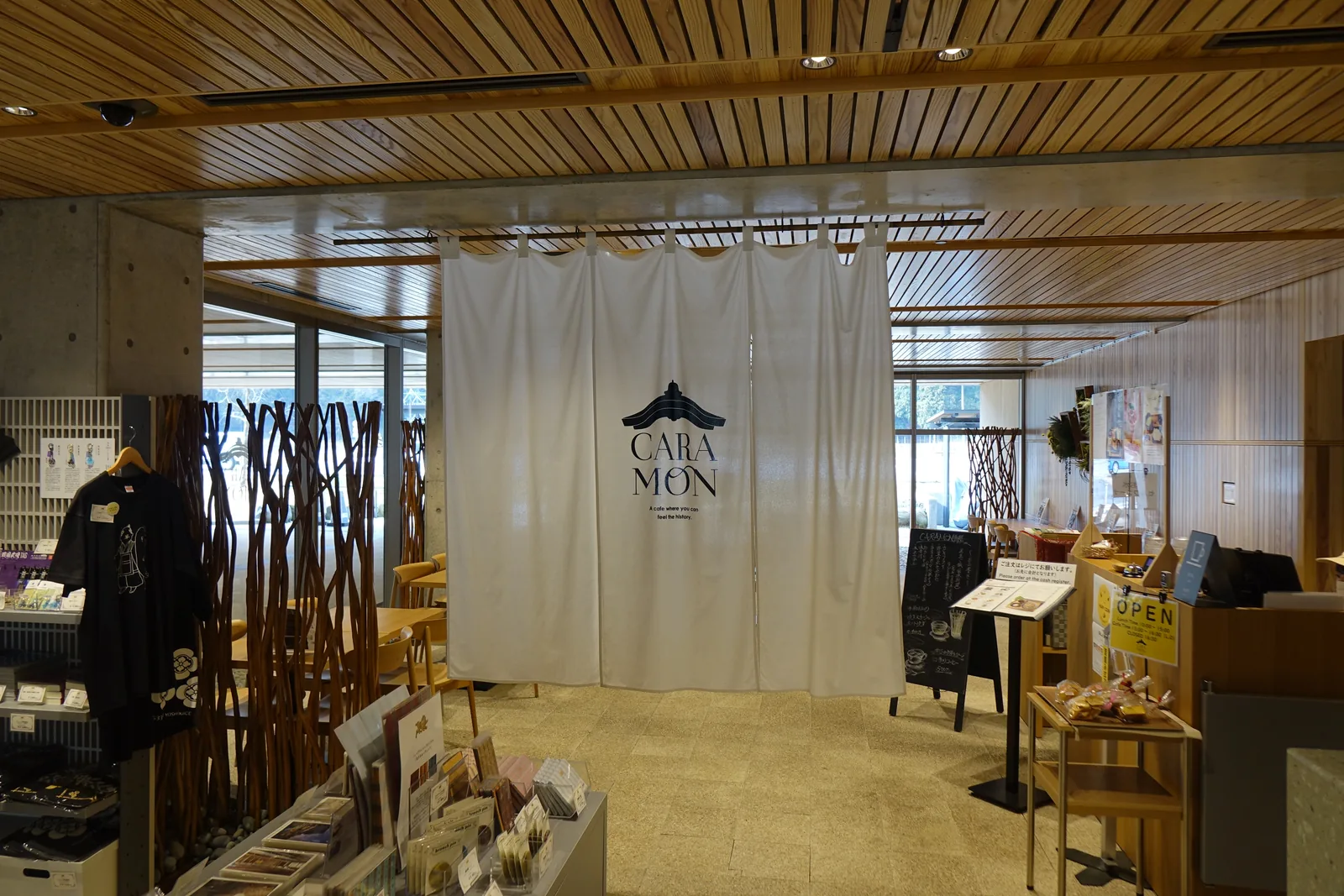 一樓大廳內的咖啡館入口，入口掛著一幅印有店名與山牆圖案的白色布簾，左側是掛著衣物與木枝裝飾的商品陳列，右側是放著商品的木架，上方是杉木板的天花