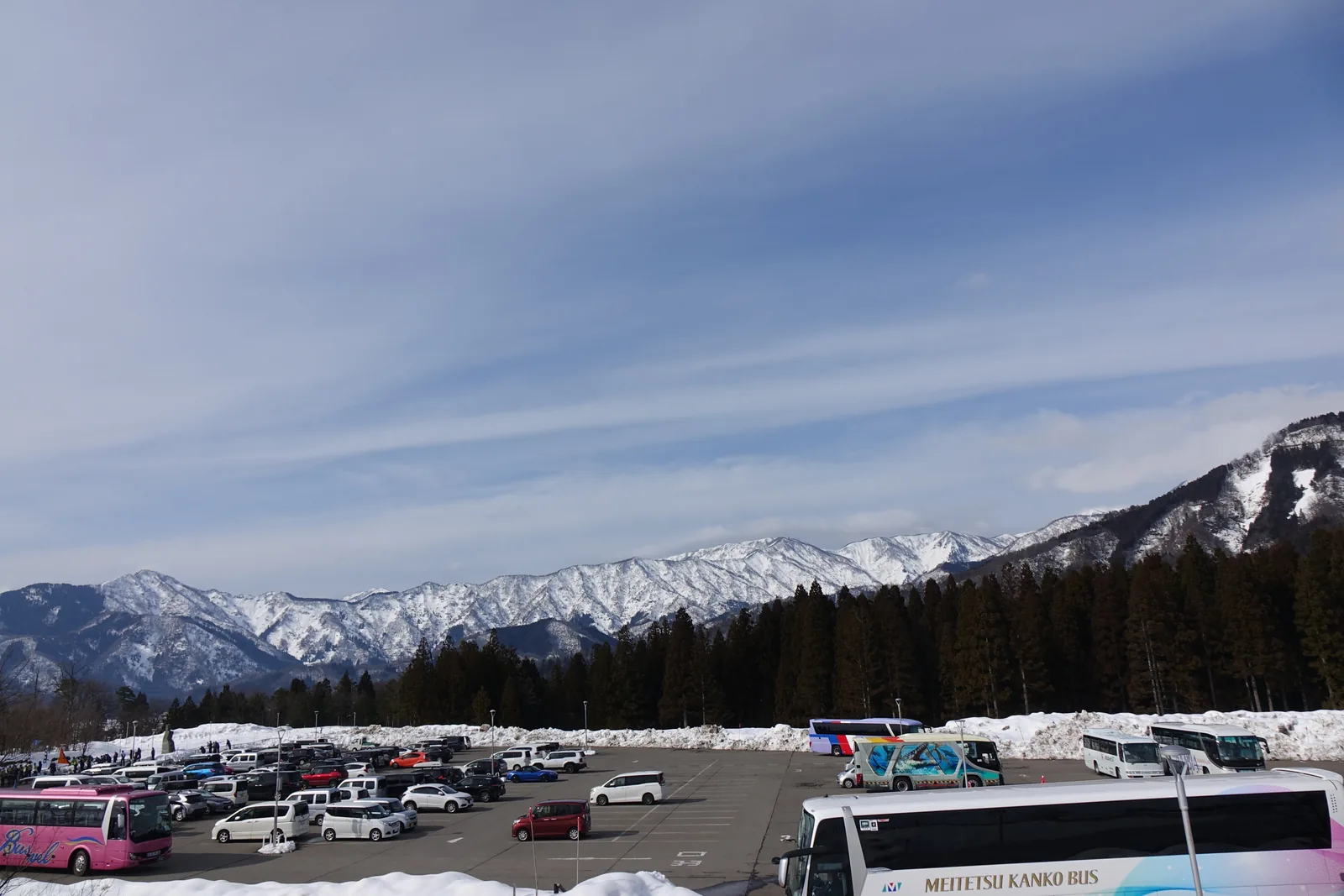 自停車場望向遠方，前景是停滿車輛的停車場與一輛大型巴士，中景是一片深色的樹林，遠處是覆著雪的連綿山稜，上方是帶著薄雲的藍天