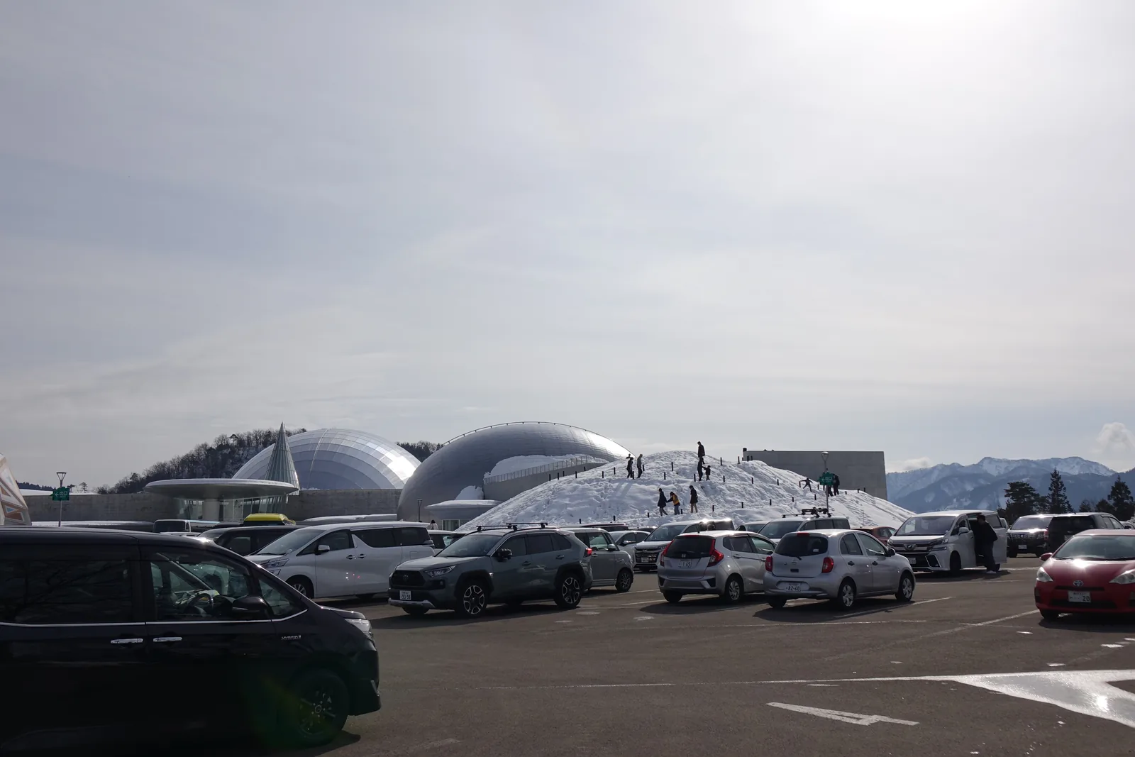 自停車場望向博物館，前景是成排的車輛，中景是一座堆高的雪丘，雪丘之後是一大一小兩座銀色的蛋形圓頂與低平的屋頂，背後是藍天