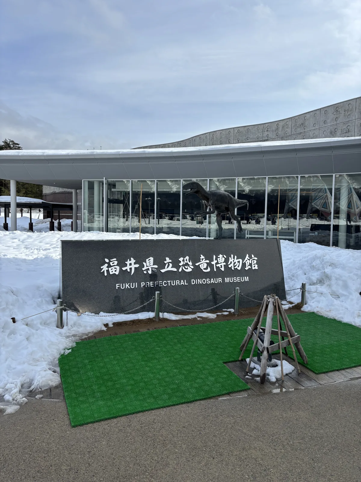 博物館的館名標示，一面深灰色的矮牆上以白字刻著館名，矮牆前鋪著綠色的地墊，背後是弧形的玻璃立面與大幅挑出的屋簷，四周是堆高的積雪