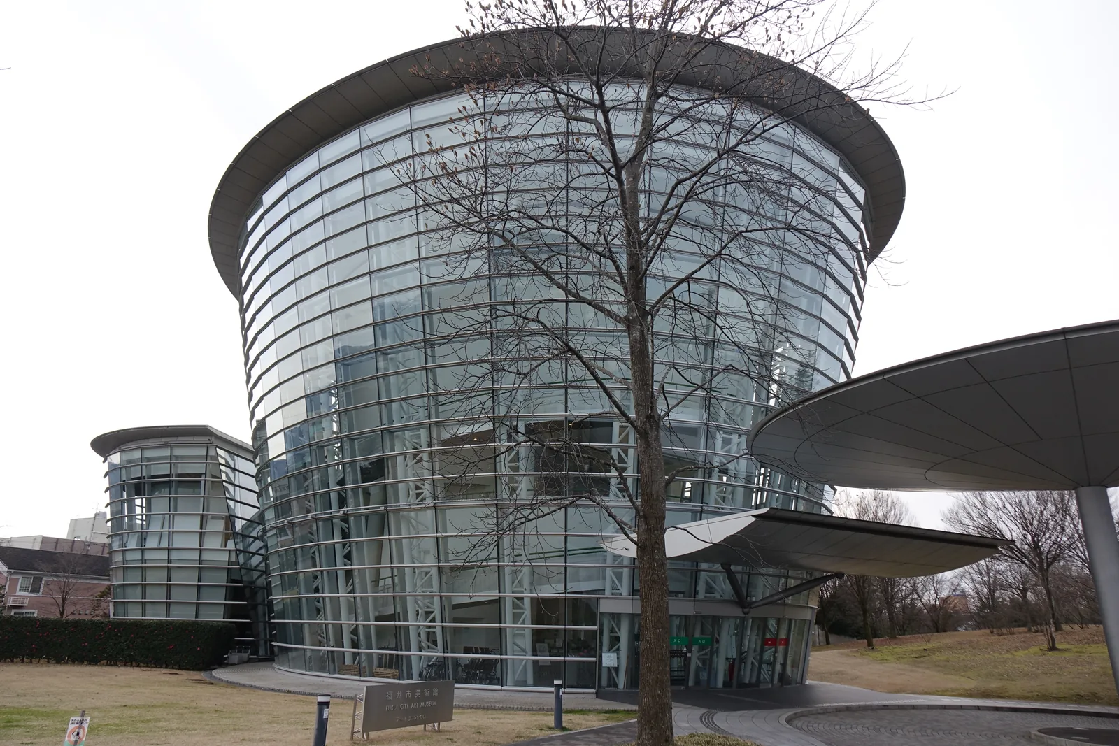公園內的一座玻璃圓筒形建築，圓筒的外牆略向內傾斜，頂部是一圈深色的寬簷，玻璃內可見白色的斜撐與樓板，圓筒的右下方伸出一片橢圓形的雨庇，前方立著一棵落葉的樹