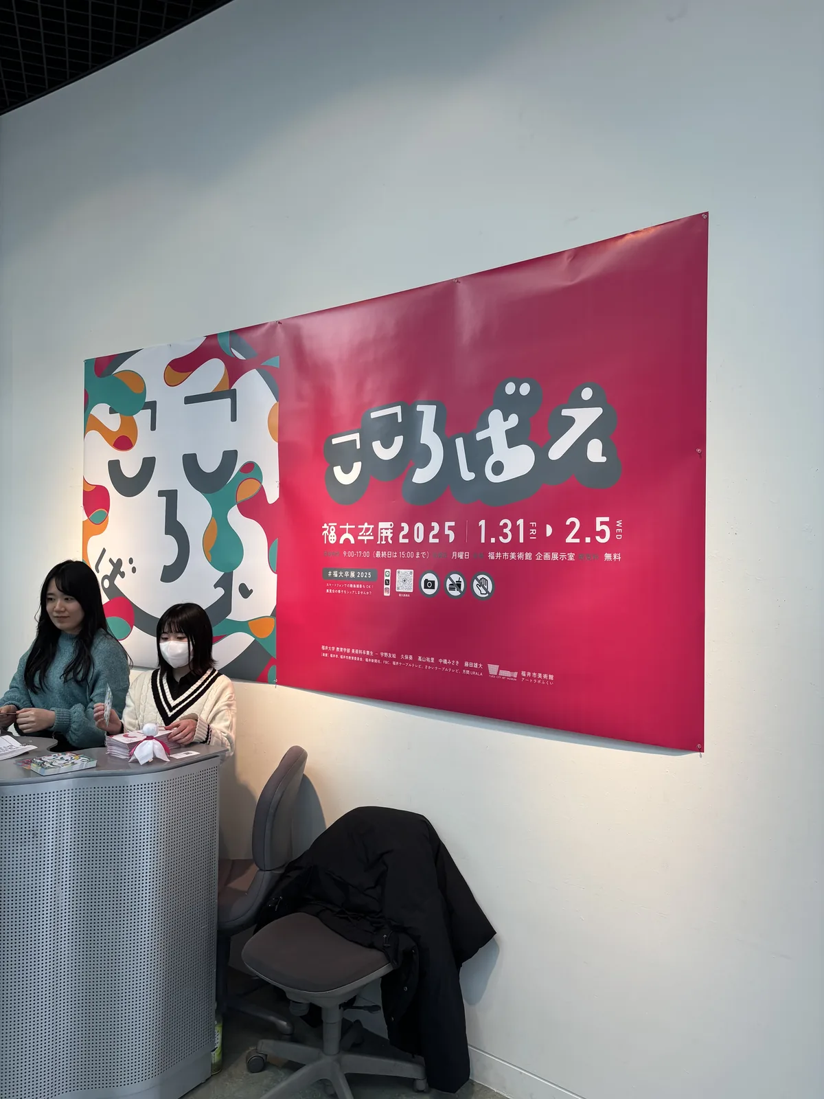 二樓展示室入口旁的受付，一面白牆上掛著一幅桃紅色的大型海報，海報上是展覽的名稱與會期，海報前是一張圓弧形的金屬色櫃台，櫃台後坐著兩位學生