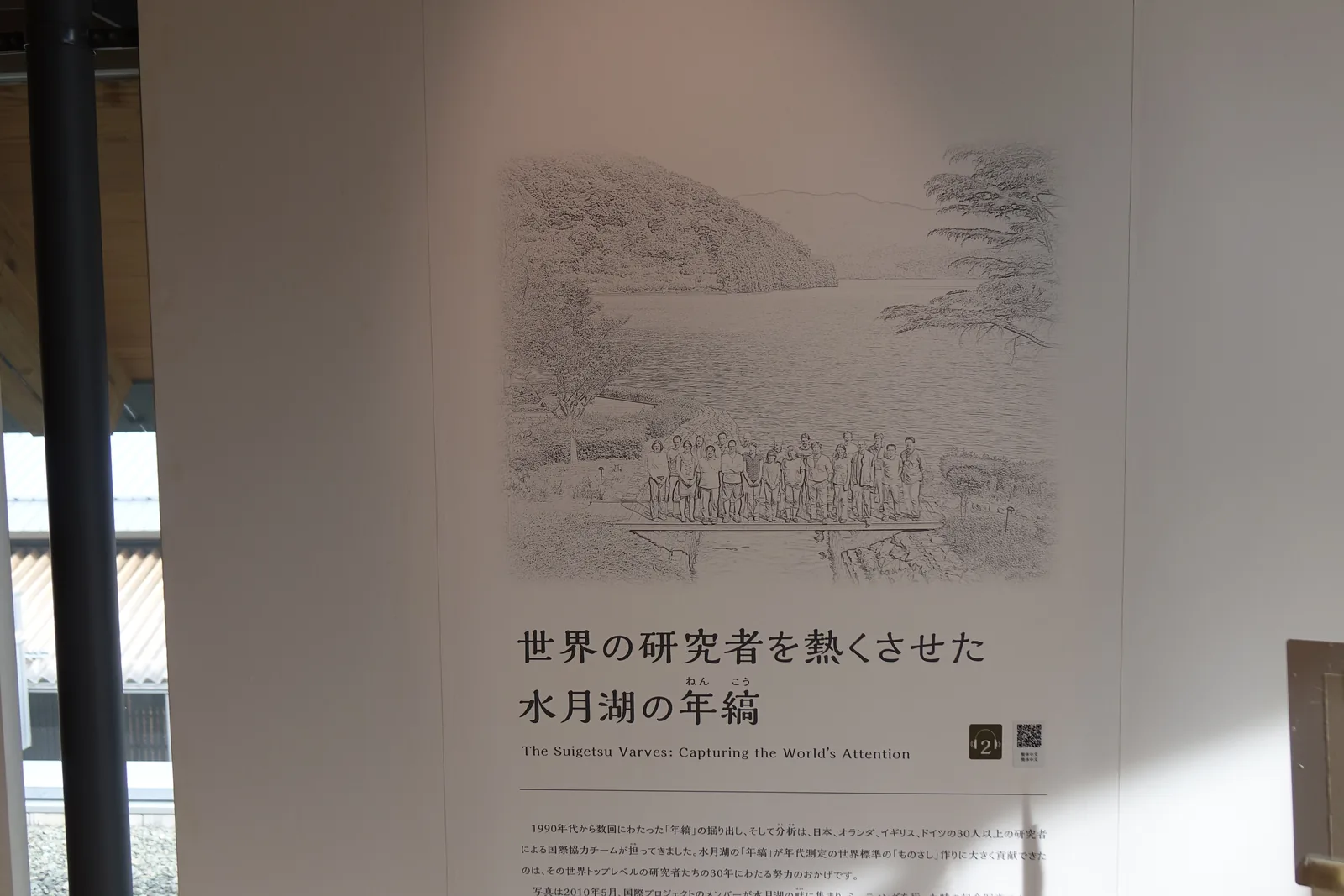 展示室入口的一面說明板，上半部是一幅以鉛筆描繪的湖與山的風景畫，下方以大字寫著水月湖的年縞令世界的研究者為之著迷，說明板立在木地板上，左側可見窗框與窗外的光