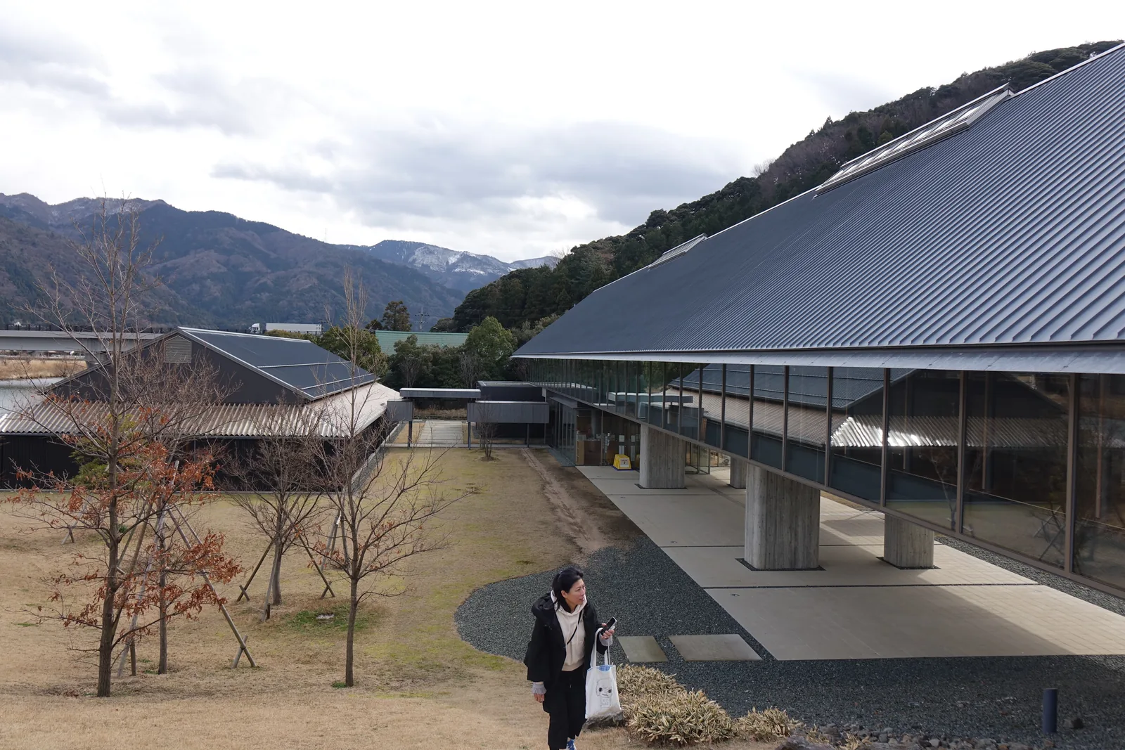 自展示棟西端的土丘望向東側，右側是展示棟的灰色斜屋頂與二樓的玻璃帶窗，一樓 piloti 的混凝土柱立在鋪石的地面上，左側是落葉的樹與一棟深色屋頂的研究棟，遠方是覆雪的山，一位參觀者自土丘走下