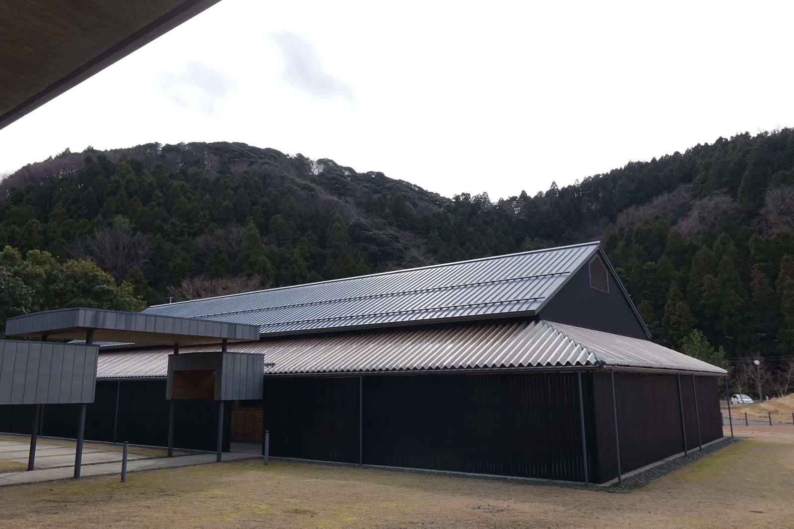 一棟黑色外牆的木造平房，切妻屋頂覆著灰色金屬板，屋頂下另有一圈較低的披屋，外牆以黑色縱向板材圍成，左側有一段金屬屋頂的連廊，背景是覆著杉林的山