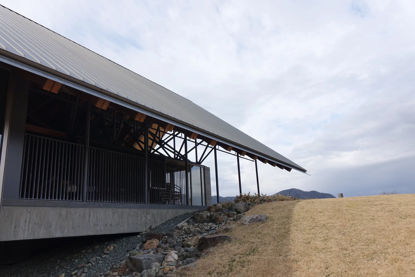 展示棟西端的屋簷，灰色金屬板的斜屋頂深深挑出，簷下可見杉木登梁的端部與斜向的鋼骨斜撐，下方是二樓的欄杆與玻璃，最下方是混凝土的樓板，樓板前方的土丘以石塊收邊，右側是枯黃的草坡