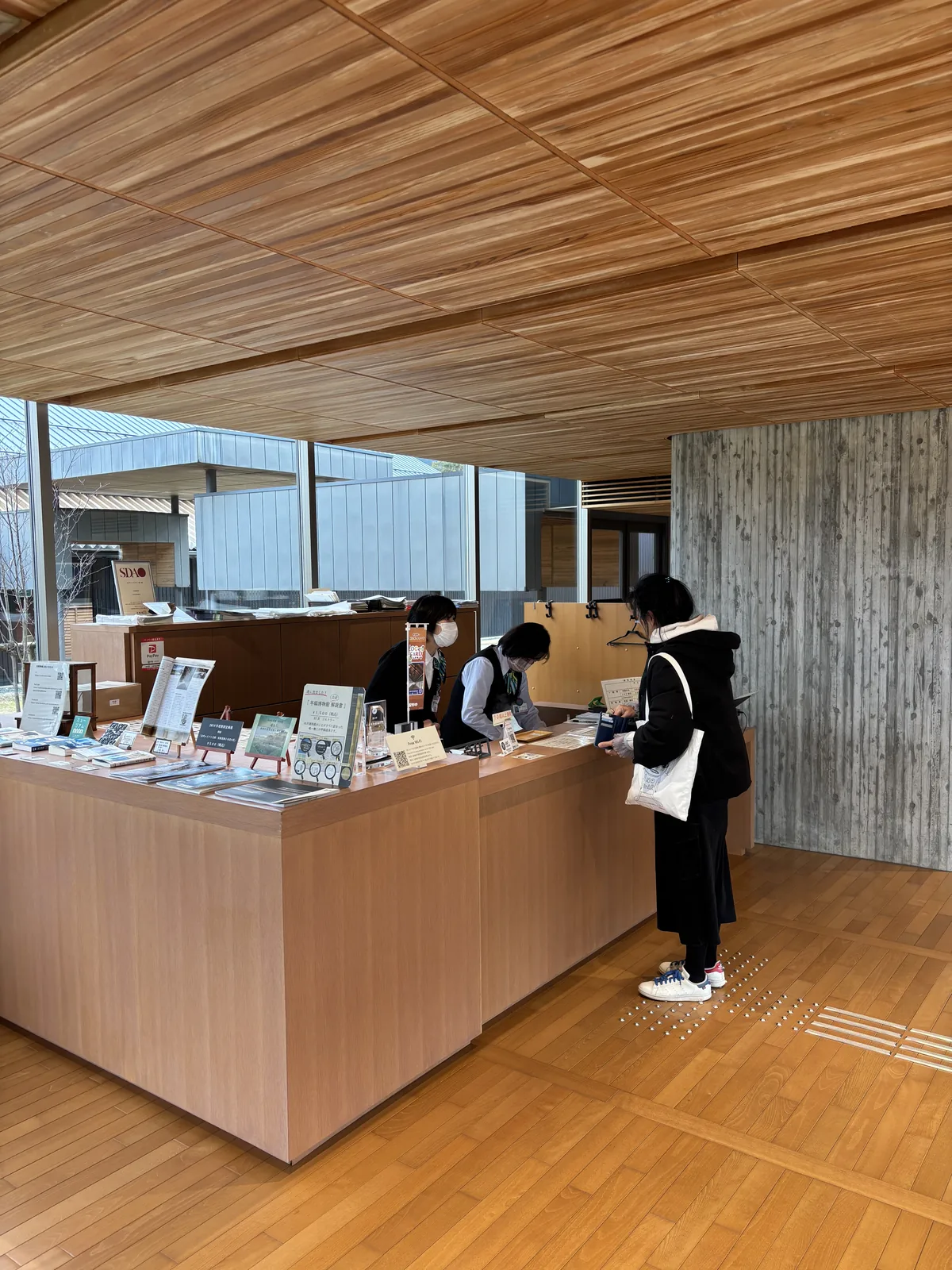 一樓大廳的受付櫃台，木製的櫃台上排著摺頁、書籍與商品，兩位職員站在櫃台後，一位參觀者在櫃台前，上方是杉木板材的天花，右後方是清水混凝土柱，左後方是大廳的玻璃與外部的景色