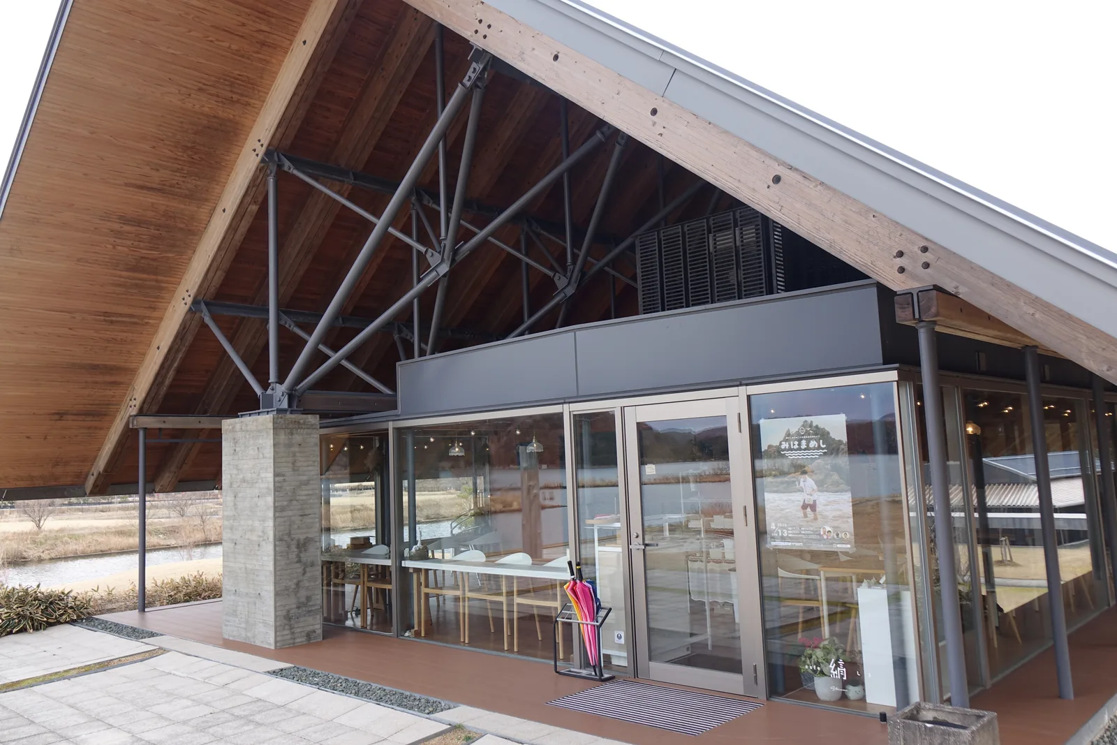 展示棟西端山牆下的咖啡館入口，上方是深深挑出的切妻屋簷，簷下可見杉木登梁與灰色的鋼骨斜材，下方是一道石材收邊的混凝土牆柱與一扇玻璃門，玻璃後方是木桌與木椅，門前是鋪石的地面