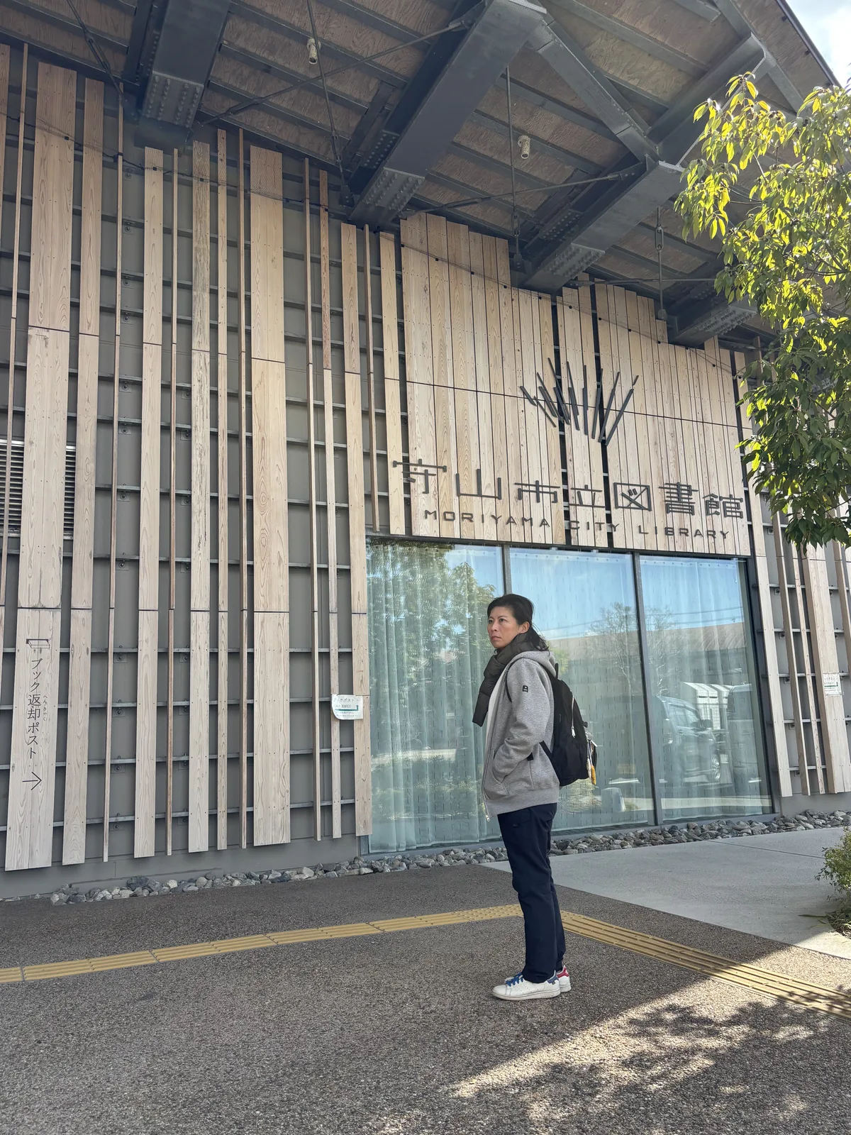 圖書館入口的外牆，上方是深深挑出的屋簷與外露的鋼構斜撐，牆面貼滿寬度不一的直向木板，木板上是館名的立體字與樹枝形的標誌，下方是一面玻璃，牆前站著一位穿灰色外套的人
