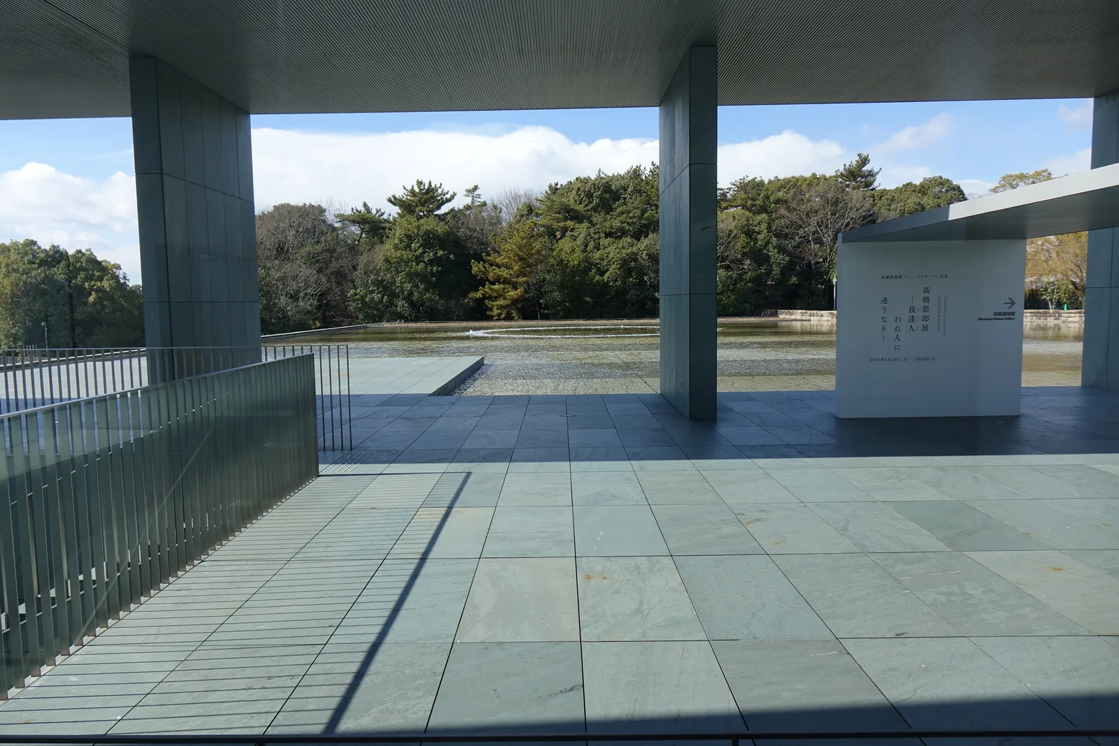 迴廊之下望向庭園，兩根灰綠色的方柱與水平的屋頂框出一片淺水池與對岸的樹林，左側是一道金屬的欄杆，右側是一面白色的說明牆，地面是灰綠色的石板