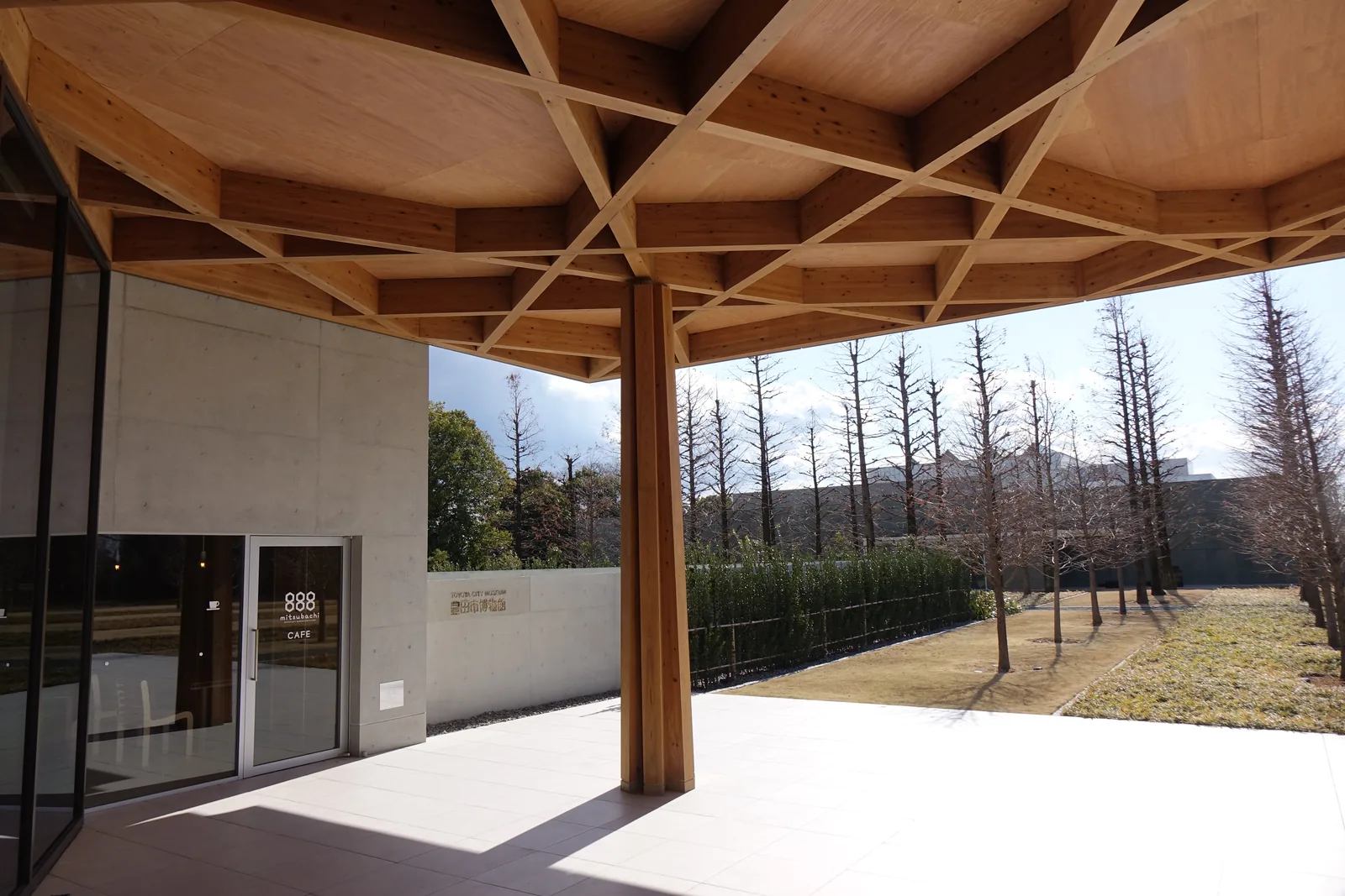 豐田市博物館的入口簷下，上方是以木材組成的格子狀屋頂，一根木柱立在淺色的地面上，左側是玻璃門與白色的牆面，簷外是成列的落葉水杉與草地