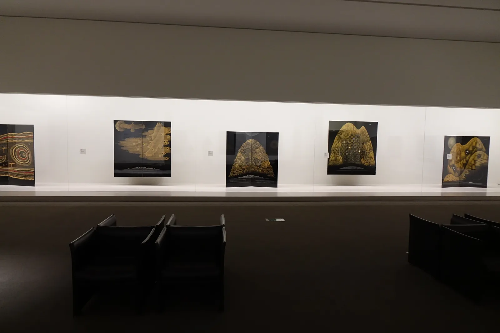 展示室的一角，深色的地毯與低矮的天花，一面白牆上排列著五件黑底金色的漆屏風作品，作品懸在牆前，前景是幾張黑色的方形座椅