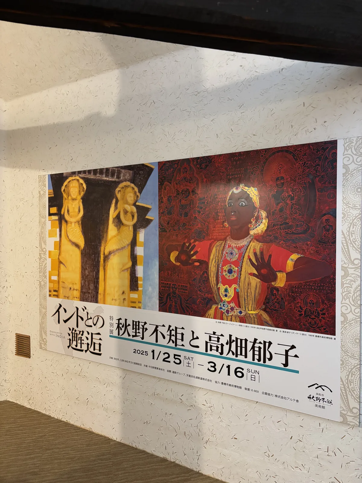 展覽海報的局部，左側是一幅以金黃色描繪的印度神像的畫作，右側是一幅以紅色為底的人物畫，下方以黑字寫著展覽名稱與會期，海報貼在一面帶有稻草纖維的白色灰泥牆上