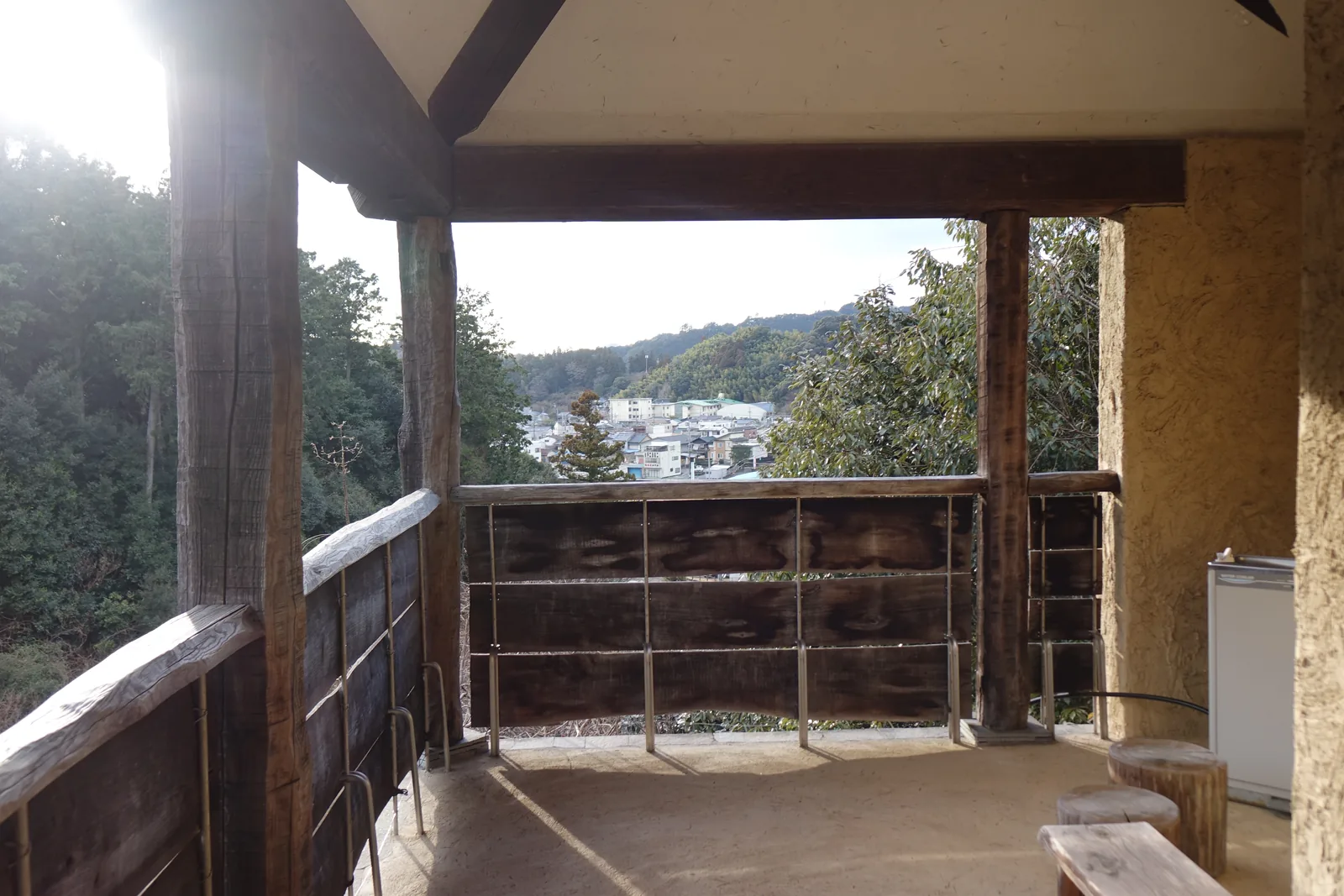 美術館二樓 terrace 的一角，深色的木柱與木欄杆框出一處開口，開口外是山谷與遠處的二俁町街區，地面鋪著木板，左側是一道土色的灰泥牆