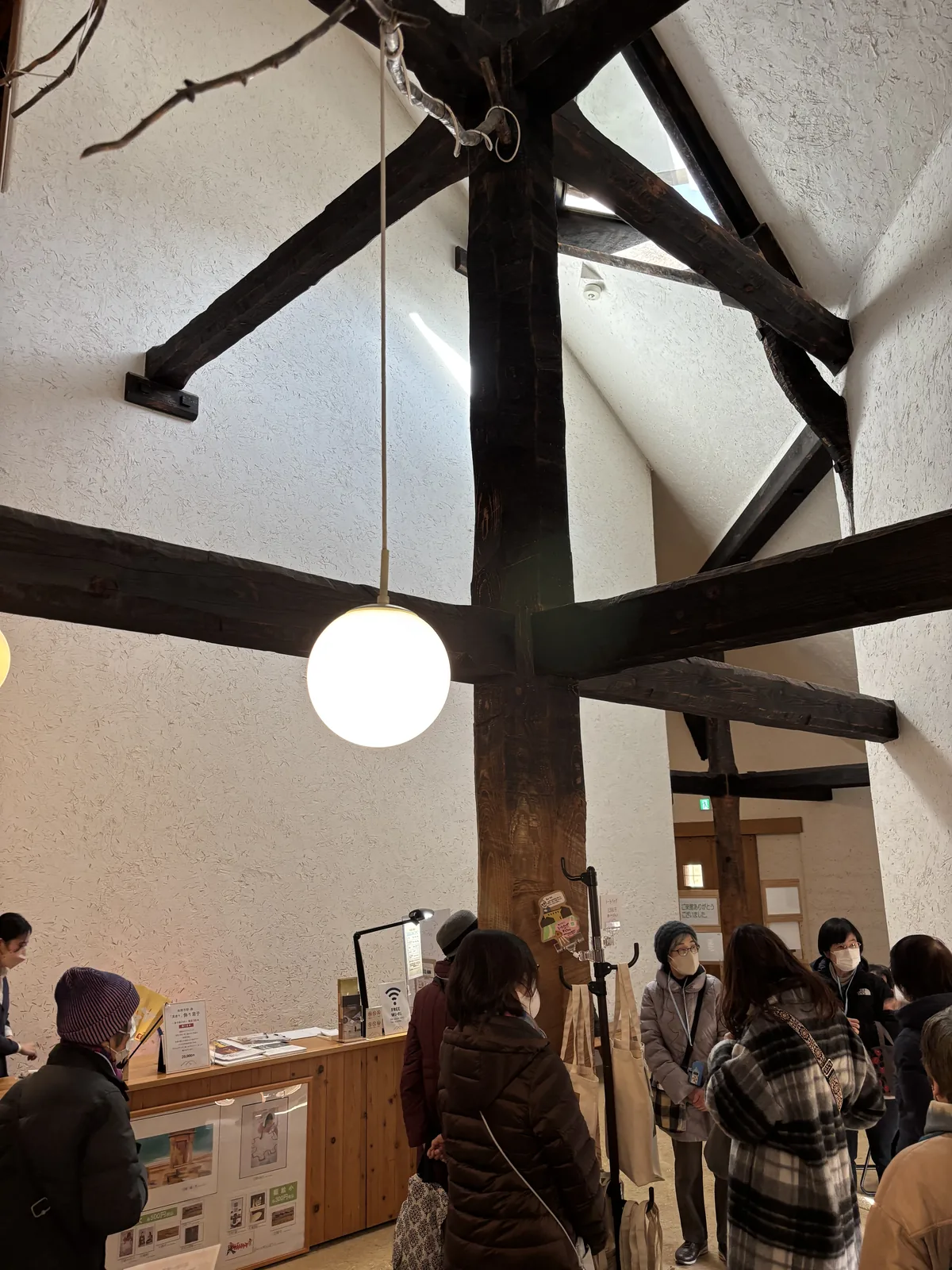 一樓的 hall，中央一根粗大的深色木柱向上延伸，柱頂有幾根斜向的深色木梁交錯，一盞球形的白色吊燈懸在梁下，牆面是白色的灰泥，下方是受付櫃台與幾位參觀者