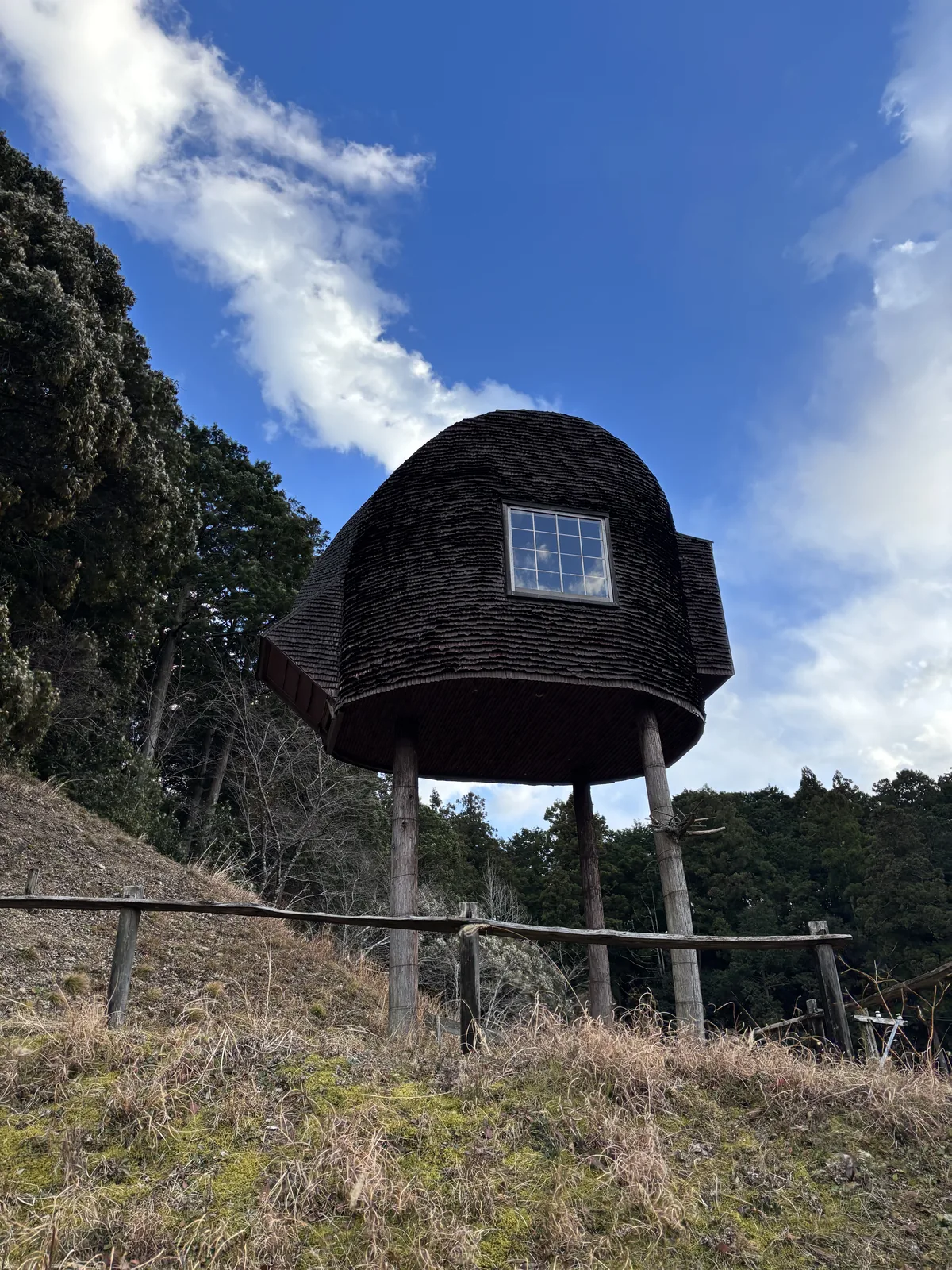 丘頂上的茶室望矩樓，一個以深色木板包覆、形如蛋形的小屋，由幾根細長的木柱腳撐起，小屋上開著一扇方窗，前景是枯黃的草坡與一道木柵，背景是藍天