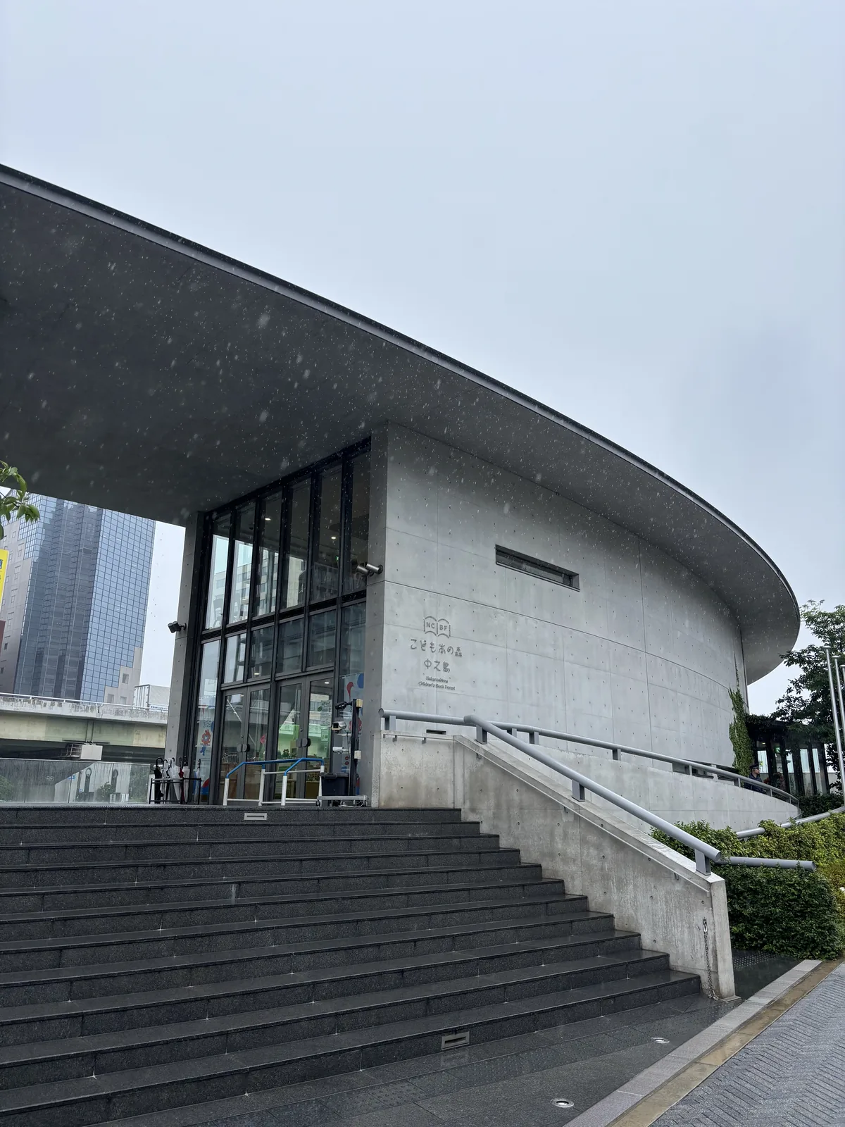雨天的圖書館入口，前景是一段深灰色的石階與斜向的混凝土扶手牆，石階之上是一片前緣呈弧線的清水混凝土大屋頂，屋頂下是嵌著小窗的混凝土牆與深色框的玻璃門
