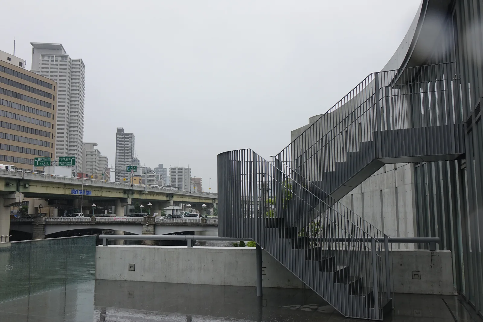 建築臨河一側的戶外樓梯，深色的鋼製樓梯與直條欄杆貼著弧形的混凝土牆折返而上，前方是混凝土的矮牆與積水的露台，遠處是高架道路與一座石造橋墩的橋