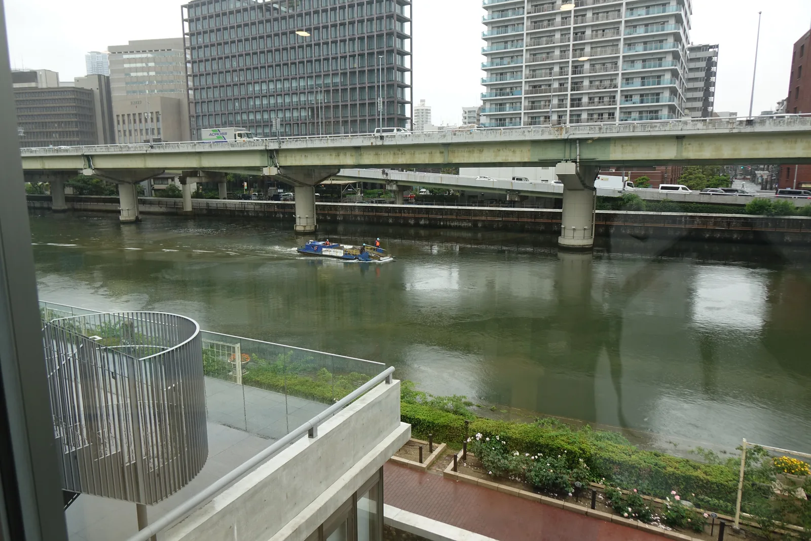 自館內的窗望向堂島川，窗外是綠色的河面與一艘藍色的船，對岸是高架道路的橋墩與成排的辦公大樓，窗下是建築的弧形露台與沿河的綠地