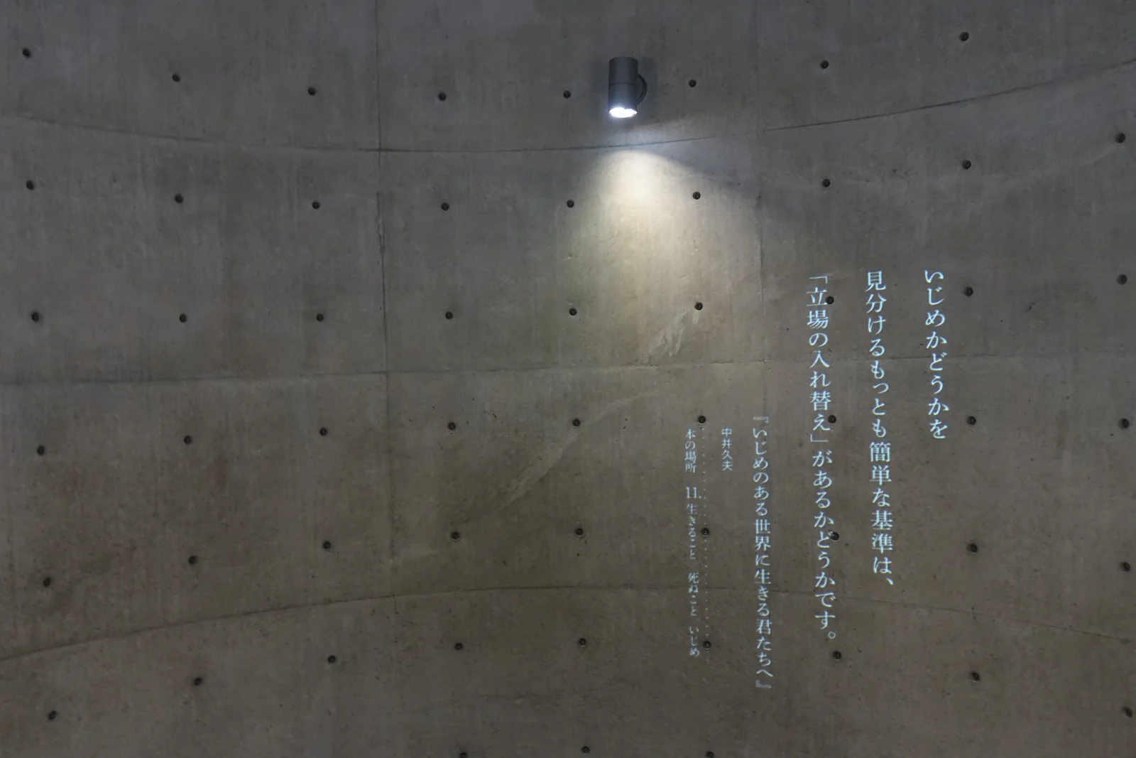 圓筒空間的筒壁，弧形的清水混凝土牆上投影著數行直書的白色文字，上方是一盞小型的壁燈