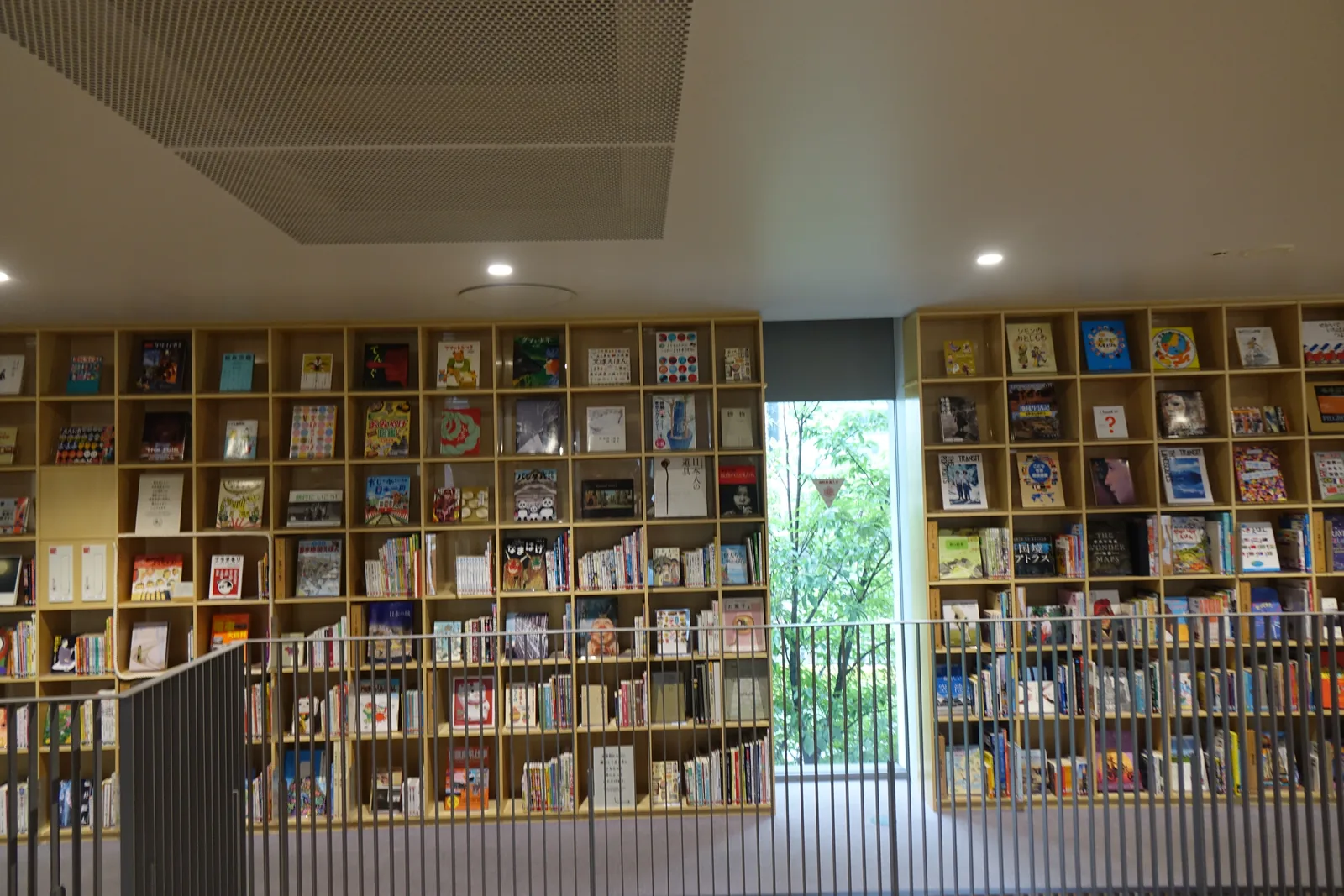 一面頂到天花的書架牆，書以封面朝外陳列，書架中央留著一道直向的細縫窗，窗外是綠樹，前方是深灰色直條欄杆的通道