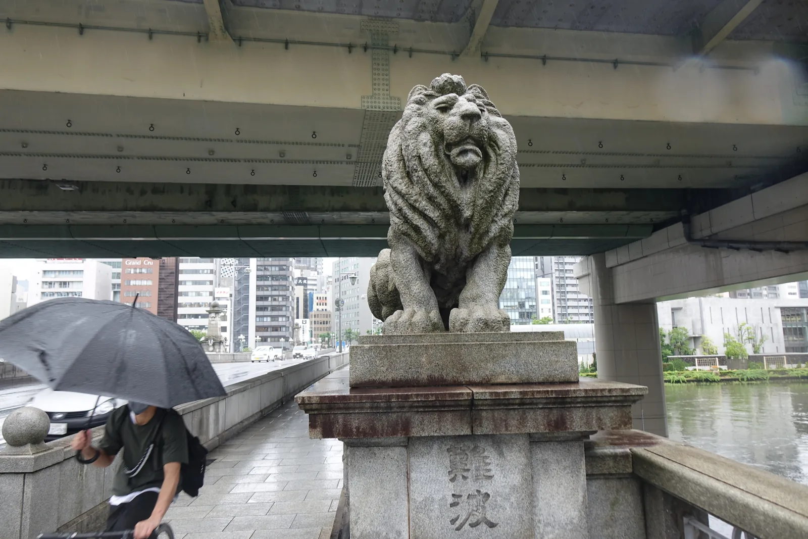 難波橋橋頭的石獅，石獅蹲坐在刻著橋名的花崗岩基座上，上方是高架道路的底面，左下方有一位撐傘的行人，遠處是河面與大樓
