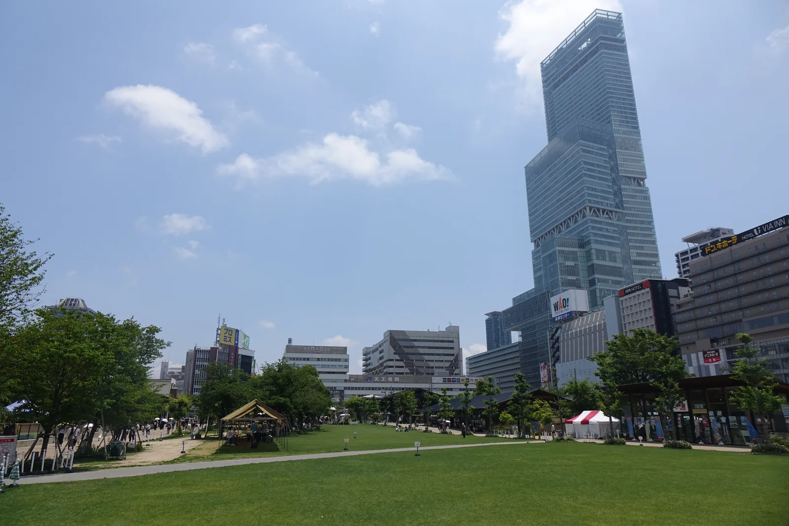 天王寺公園的草坪廣場，前景是大片的草坪，草坪的邊緣是成排的低矮木造店舖與樹，右後方是一棟玻璃帷幕的超高層大樓與車站一帶的建築