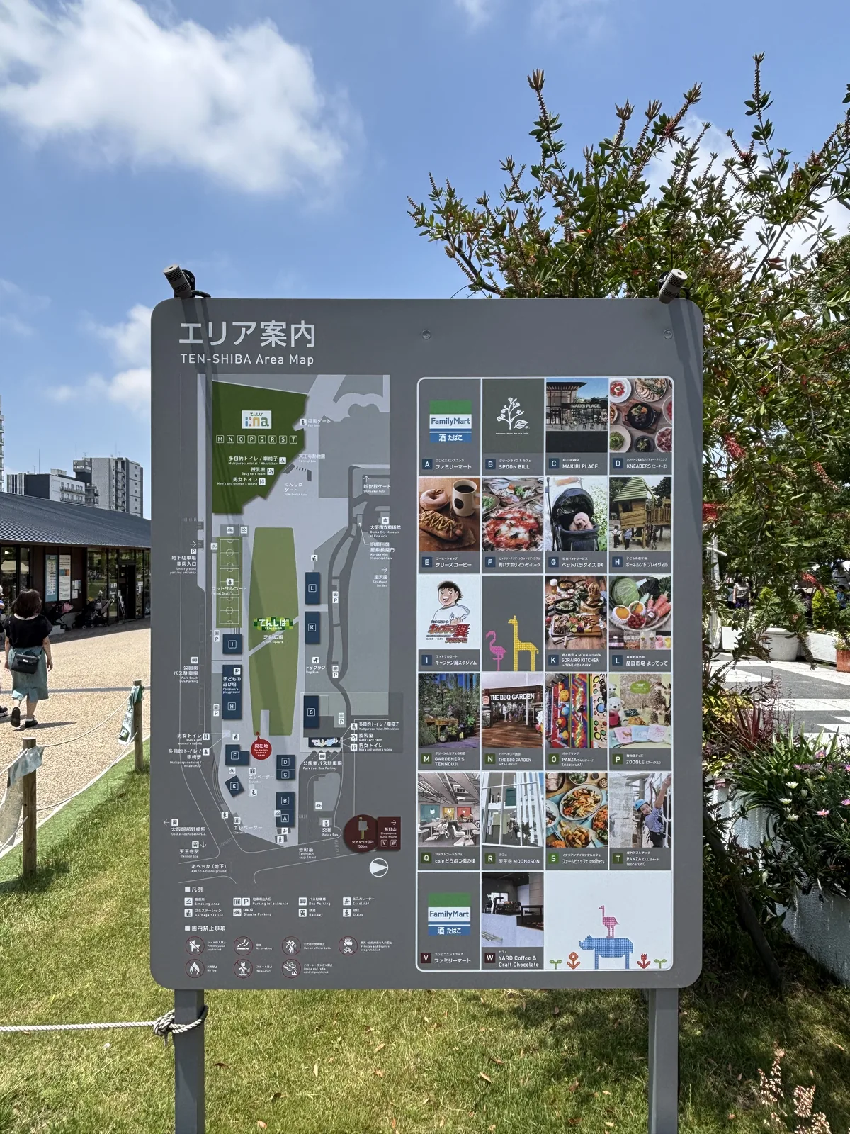 公園入口區的案內板，板面上是公園的平面圖與各店舖的照片，案內板立在草地與步道之間，後方是店舖與樹